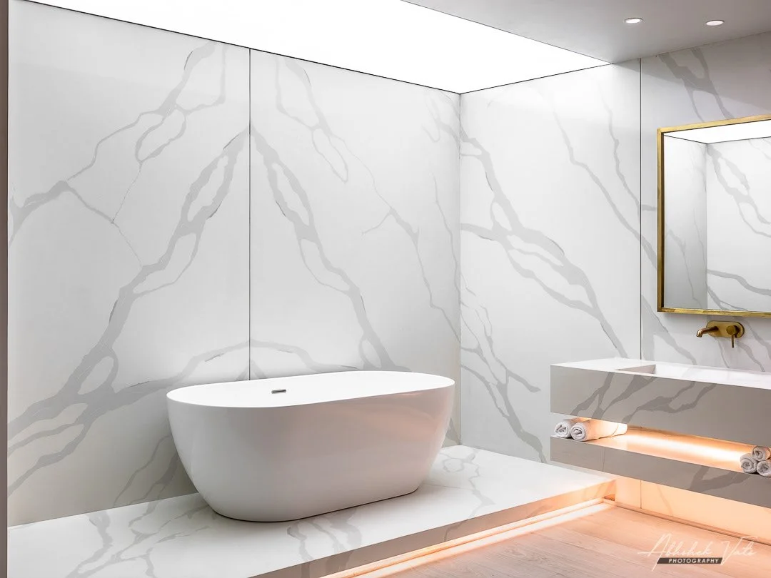 Modern bathroom with white marble walls and floor, minimalist bathtub, wall-mounted sink with rolled towels and ambient lighting.