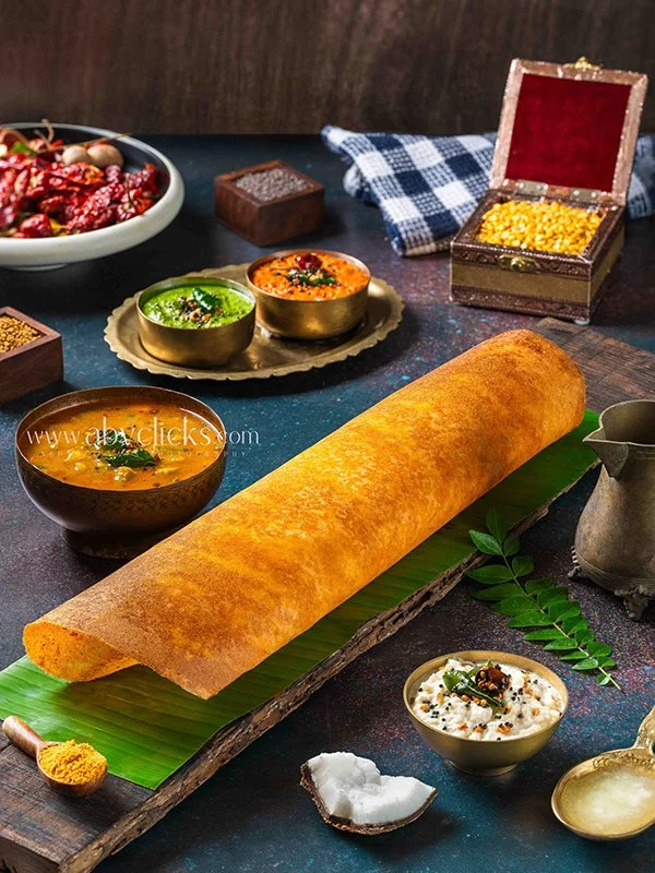 A large crispy dosa rolled on a banana leaf, with bowls of chutneys, sambar, and various Indian condiments around it. There is a tray with colorful side dishes and a metal jar, on a dark surface with a checkered cloth in the background.