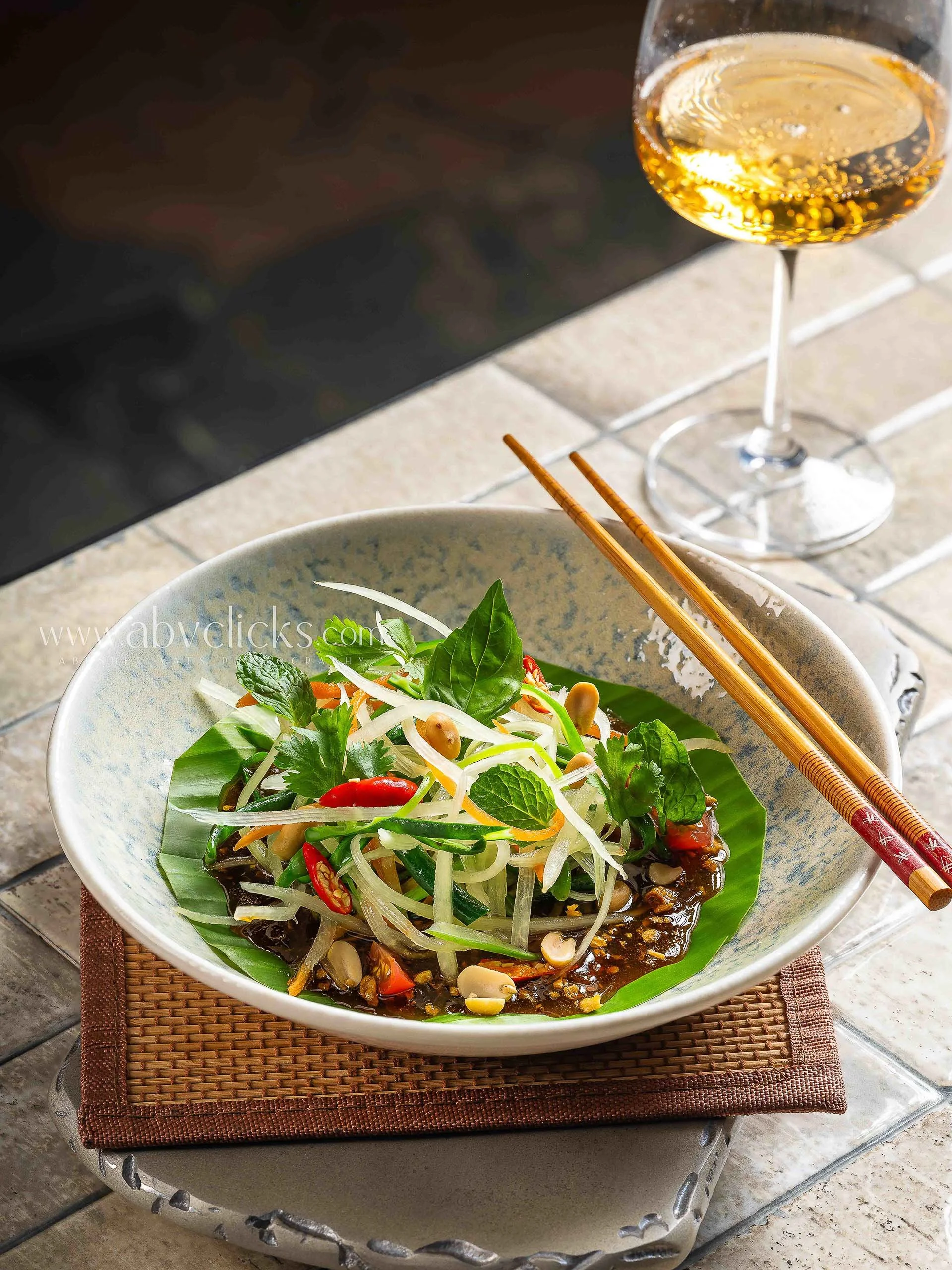 A bowl of salad with fresh greens, sliced vegetables, and herbs, placed on a woven mat with chopsticks resting on the bowl. In the background, there is a glass of sparkling wine or champagne.