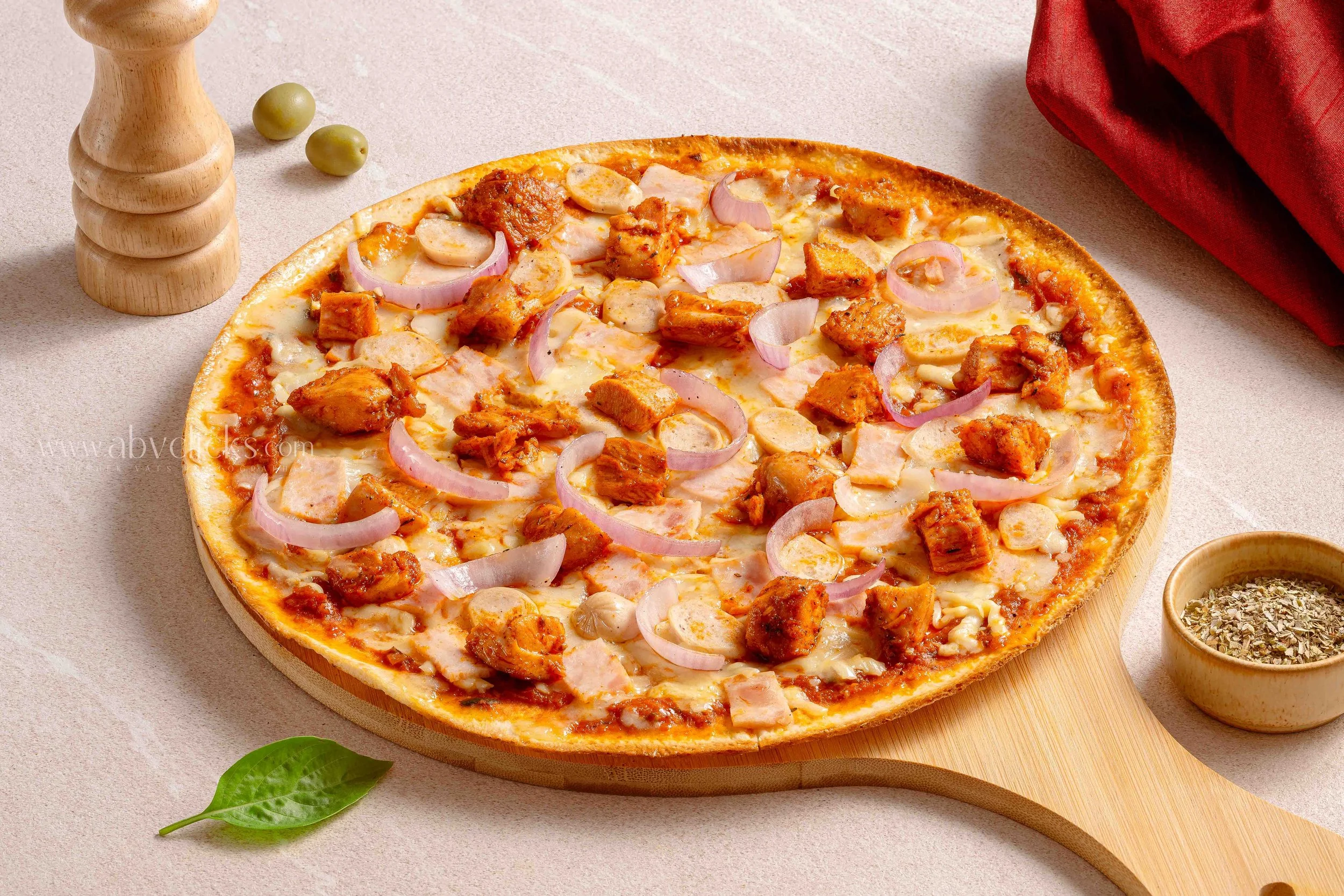 A pizza with chicken, onions, and cheese on a round wooden board, accompanied by small bowls of herbs and spices, two green olives, and a red cloth.