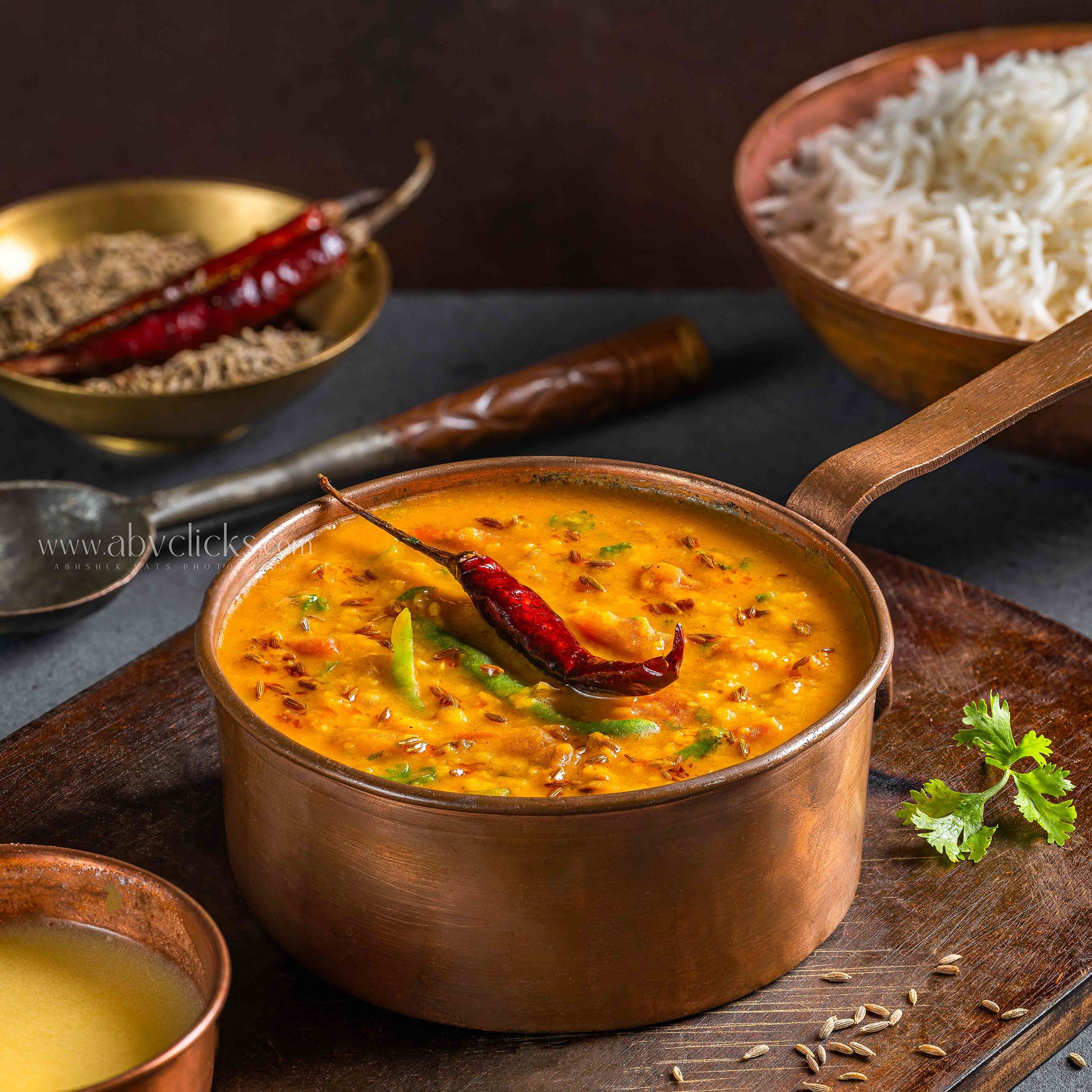 A copper pot filled with a yellow lentil soup garnished with green herbs and a dried red chili on top, placed on a dark wooden surface with scattered cumin seeds and a sprig of cilantro nearby. In the background, there are bowls containing rice and s
