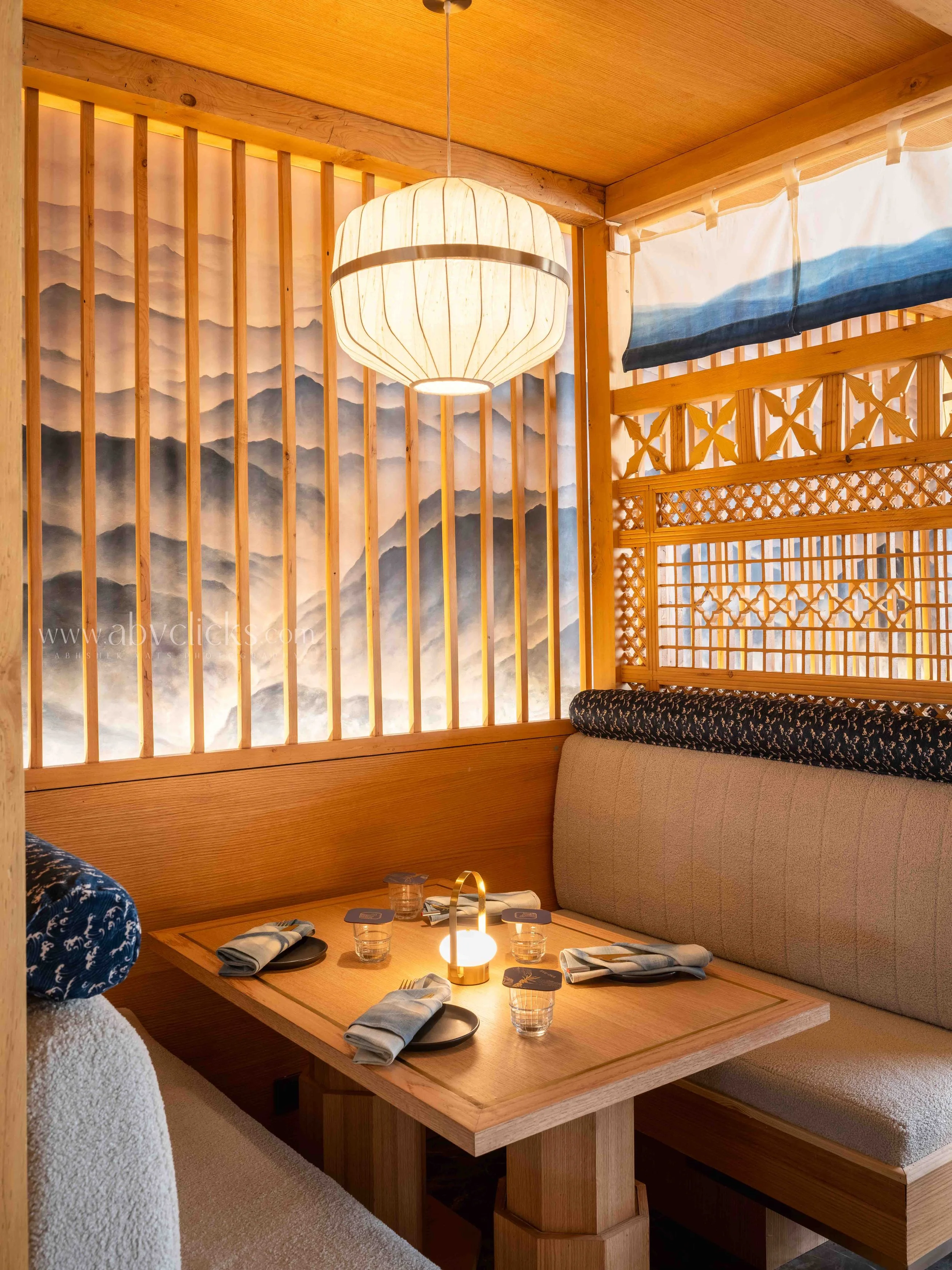 A cozy restaurant booth with wooden dividers and a scenic mountain-themed wallpaper, illuminated by a paper lantern overhead, with a table set with glasses, napkins, and utensils.