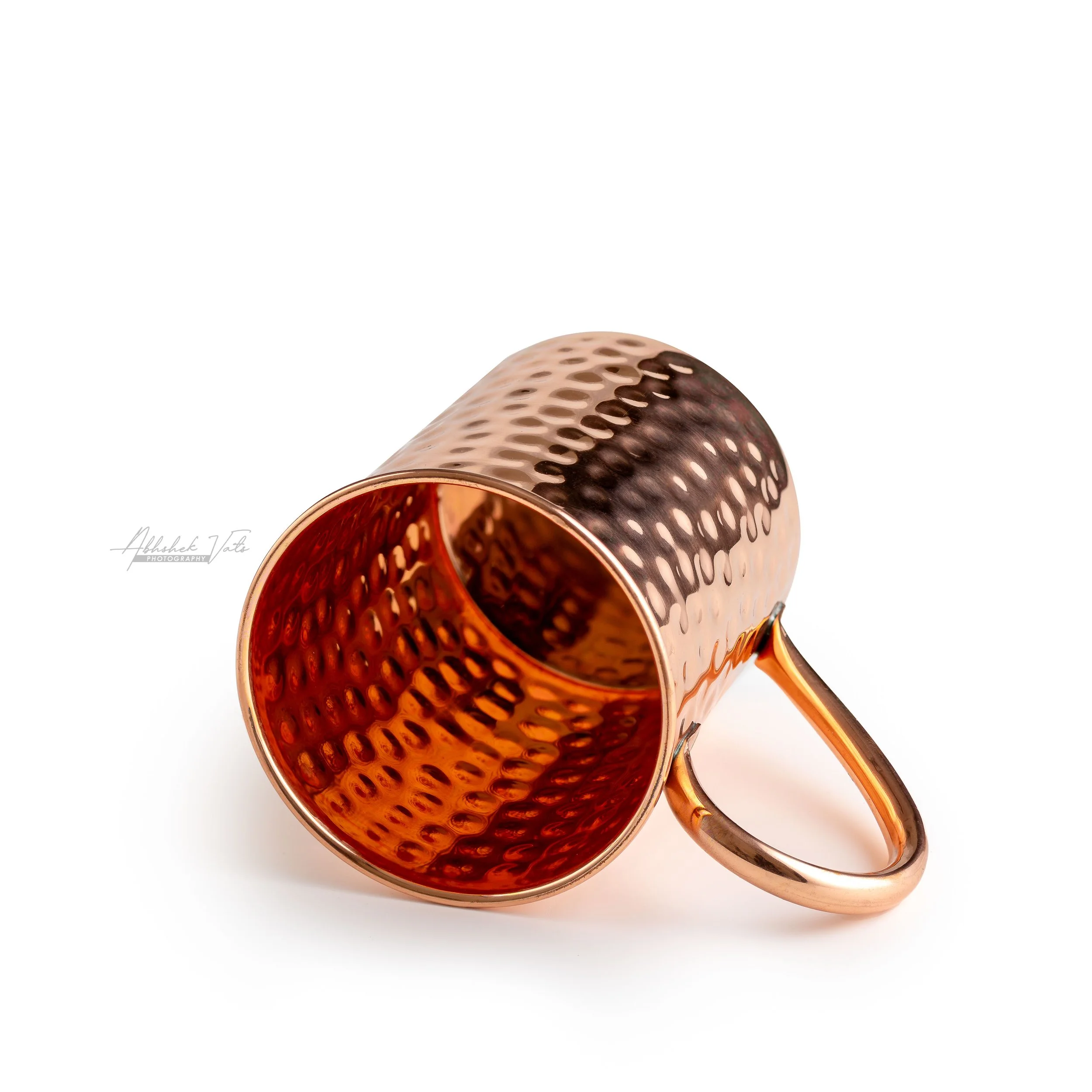A close-up of a copper-colored, hollow, perforated metallic mug or cup with a handle, laying on its side on a white surface.