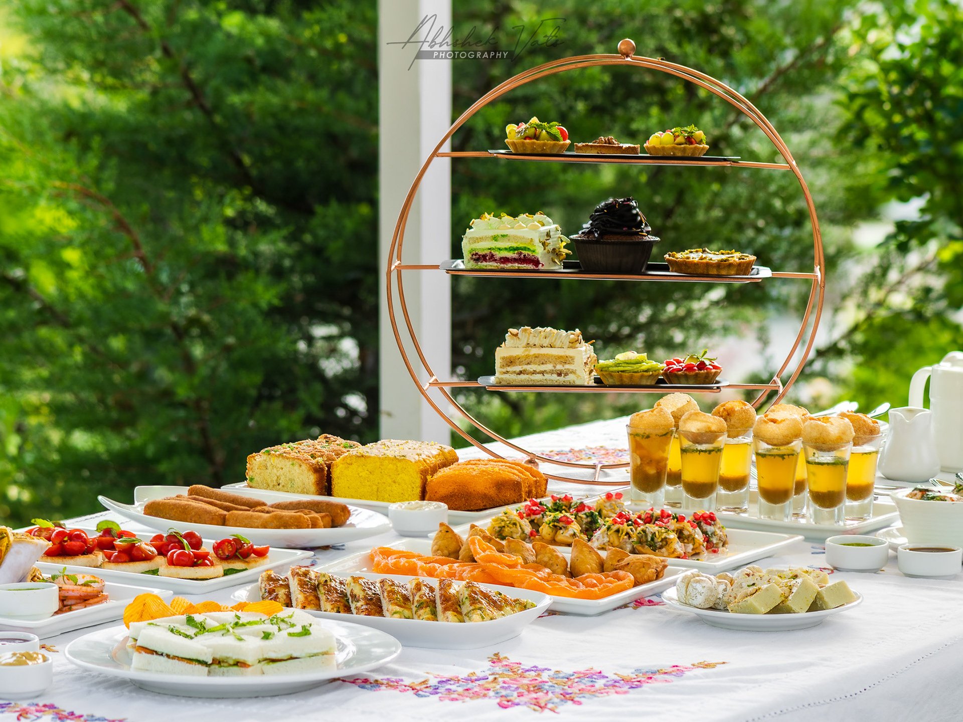 A large outdoor table filled with a variety of desserts, appetizers, and drinks, with a three-tiered dessert stand and sunlight filtering through green trees in the background.