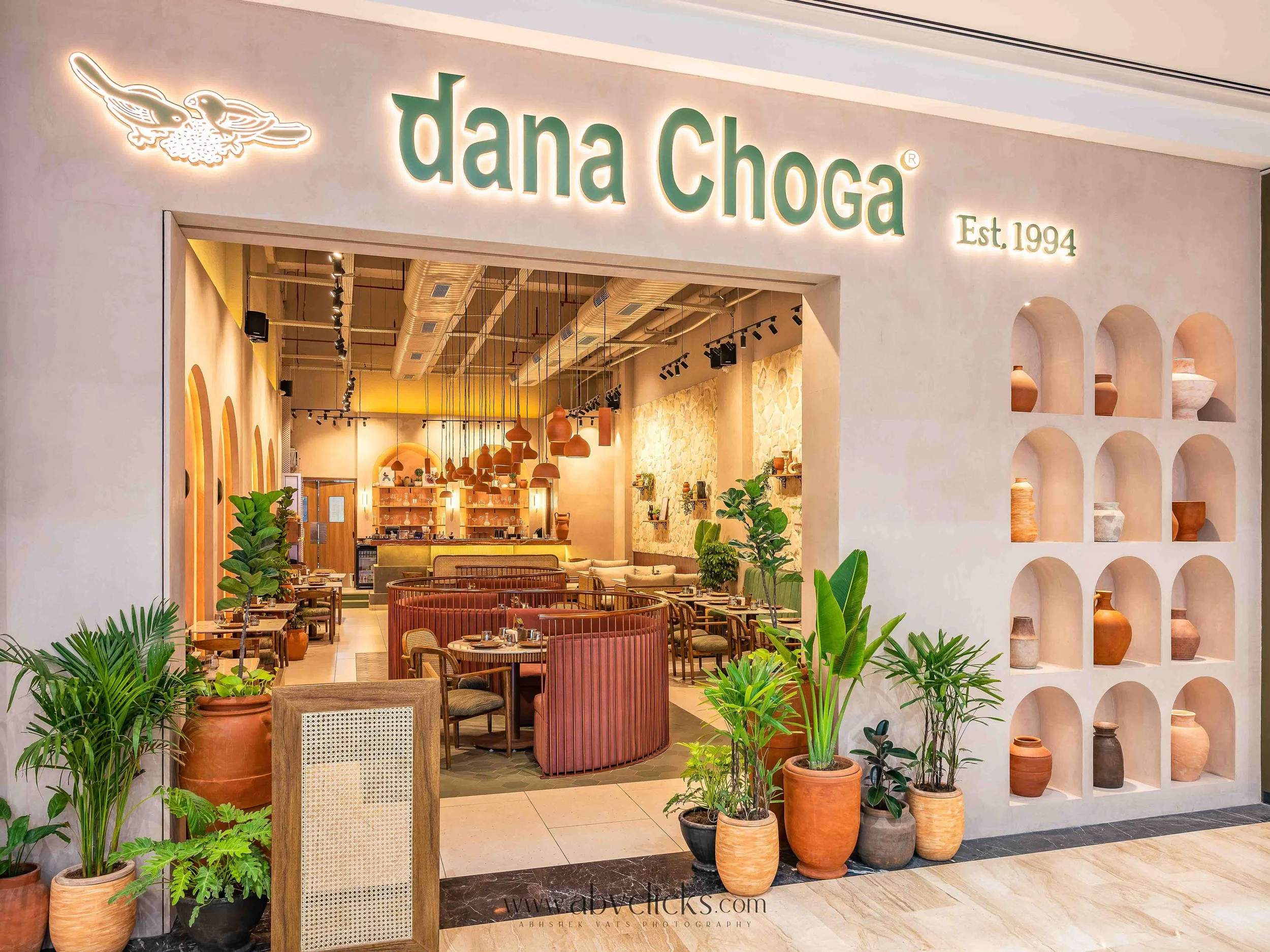 Storefront with sign 'Dana Choga' and 'Est. 1994' on a light-colored wall. Inside, there are warm-toned hanging lamps and a cozy dining area with plants and various decorative pots on display.