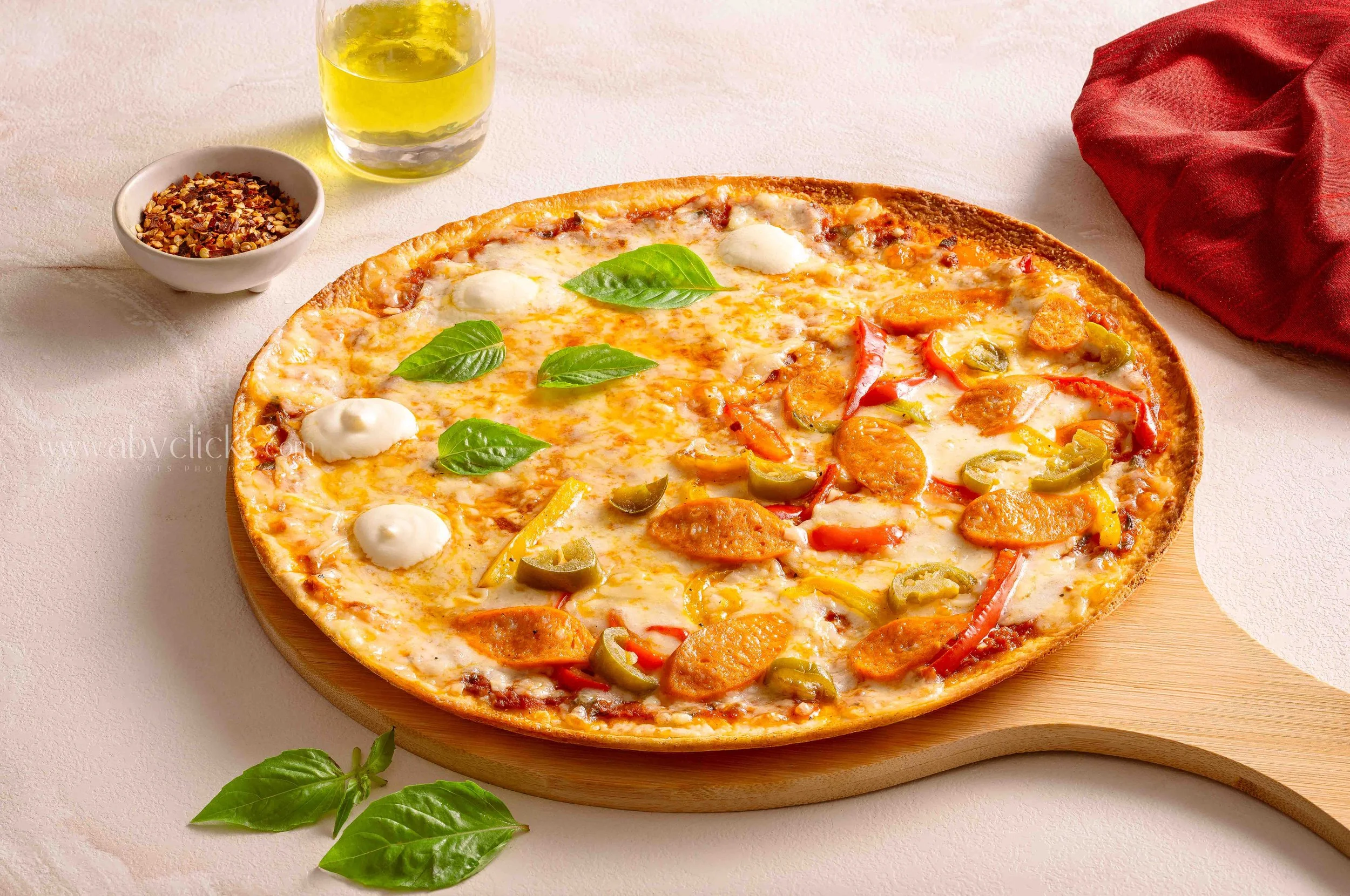 A cheese pizza topped with slices of pepperoni, green bell peppers, and red peppers, garnished with basil leaves. Accompanied by a small bowl of red pepper flakes, a glass of olive oil, and a sprig of basil on a white surface.