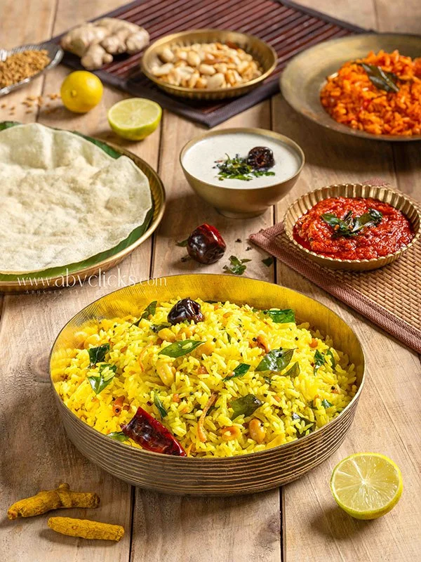 A bowl of yellow rice garnished with herbs, surrounded by various Indian dishes and condiments on a wooden table.