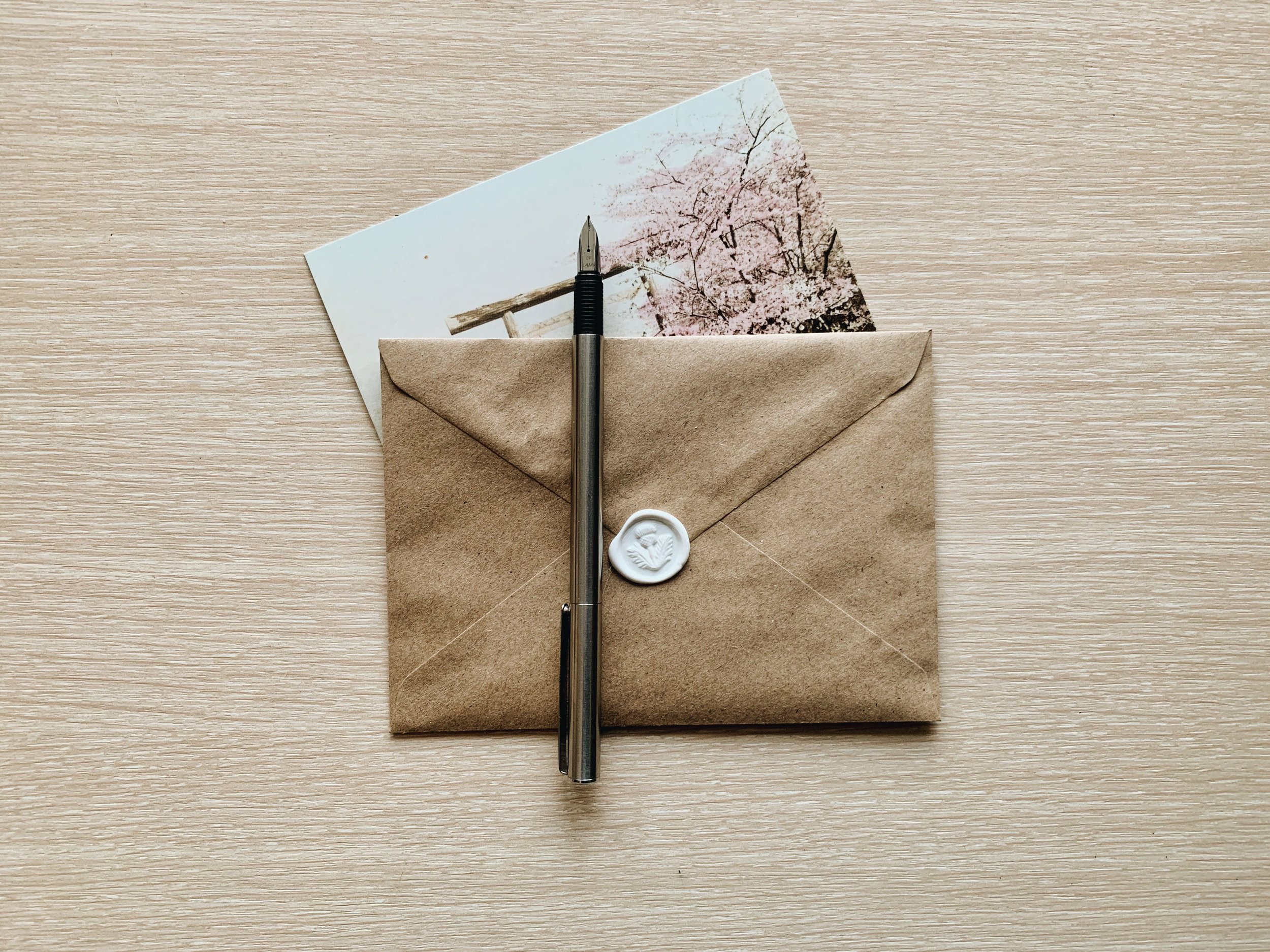 A brown envelope with a white wax seal, a pen, a postcard with cherry blossoms, and a bamboo stick on a light wooden surface.