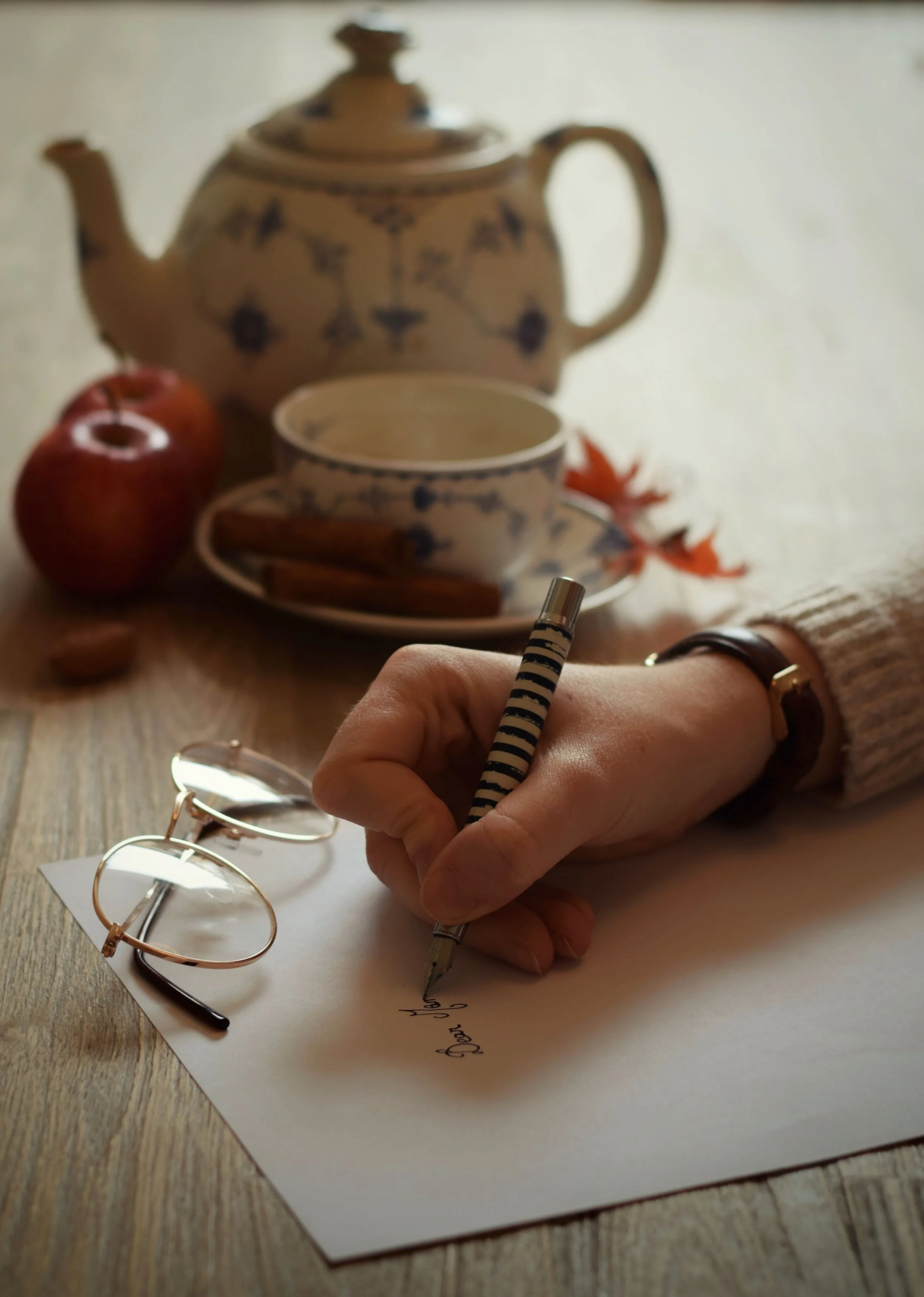 A person is writing a note on a piece of white paper, holding a striped pen. The person is wearing a watch and several rings. On the wooden surface near the paper, there are eyeglasses with round frames. In the background, there is a ceramic teapot with a floral pattern, a matching teacup, a cinnamon stick, a red apple, and some red leaves.