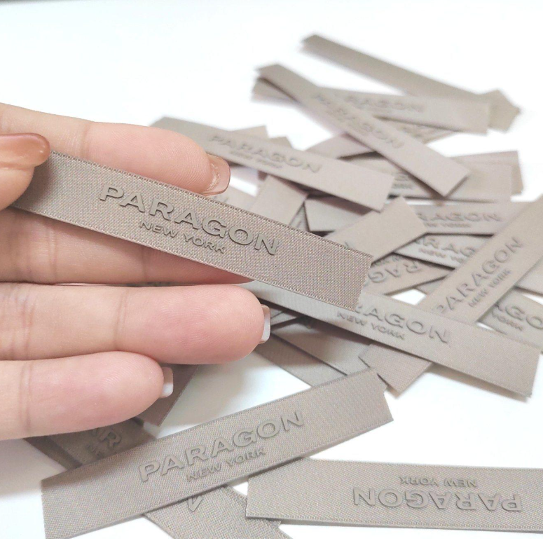 A hand holding a beige fabric label with the words 'PARAGON NEW YORK' embossed on it, surrounded by many similar labels scattered on a white surface.