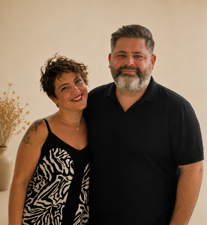 A woman with short, curly hair and a tattoo on her shoulder, wearing a black and white patterned dress, standing next to a man with short, gray hair and a beard, wearing a black polo shirt, both smiling and standing close together.