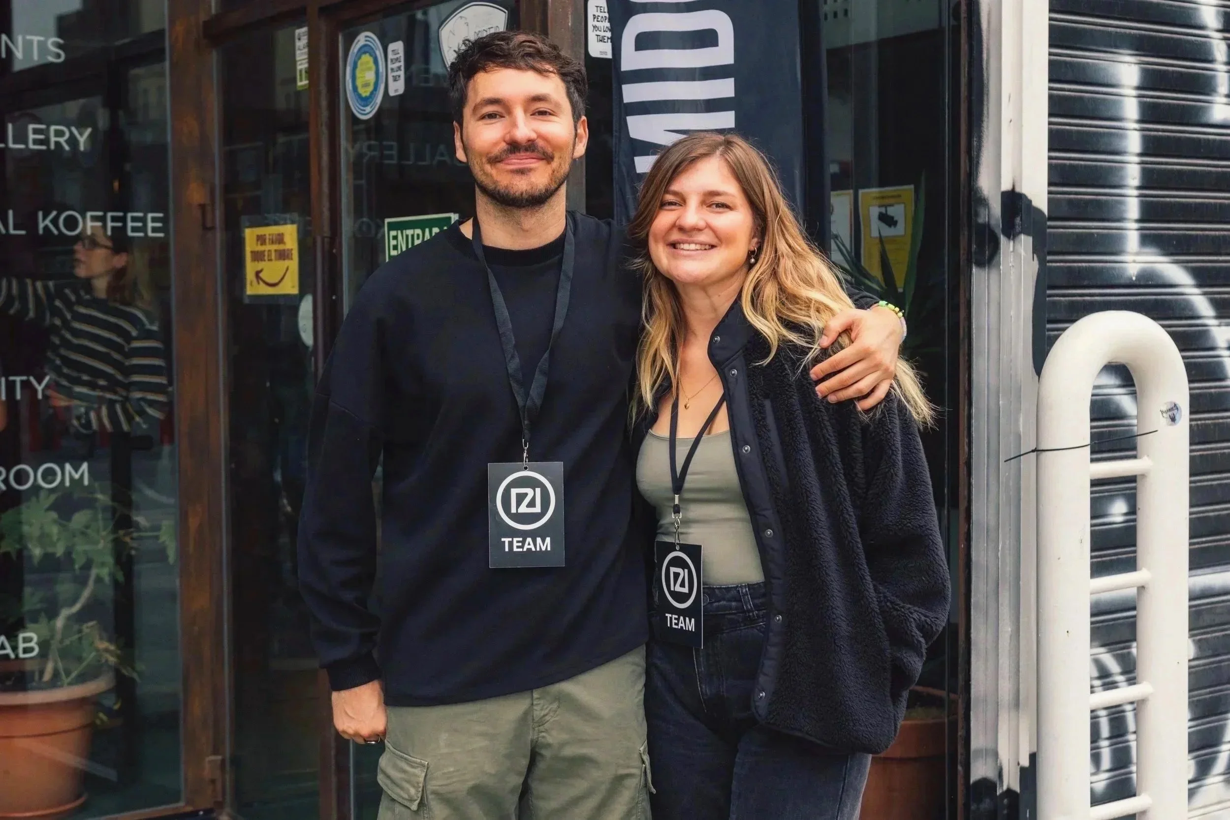 A man and a woman smiling and hugging outdoors in front of a shop entrance, both wearing black lanyards with badges that say 'TEAM' and the number 12.