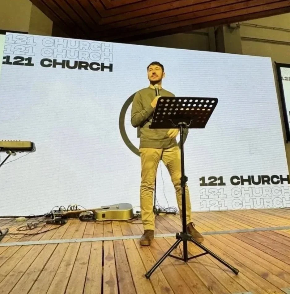 A man speaking into a microphone on stage at 121 Church, with musical equipment around and a large screen behind him displaying the church's name.