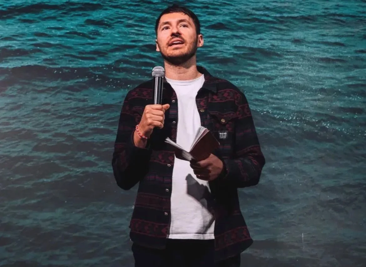 A man holding a microphone and a notebook, speaking in front of a water-themed background.