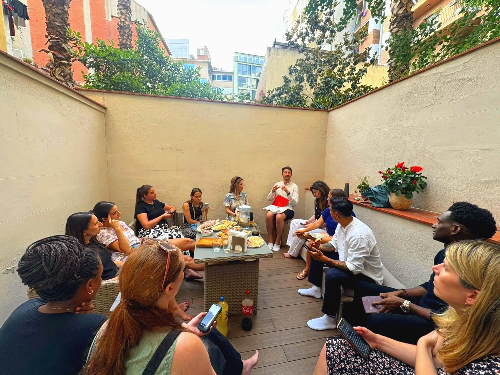 A group of people gathered on a small outdoor patio, sitting on chairs and benches, participating in a social event or meeting. There are various snacks and drinks on a table in the center, and the atmosphere appears lively and engaged.