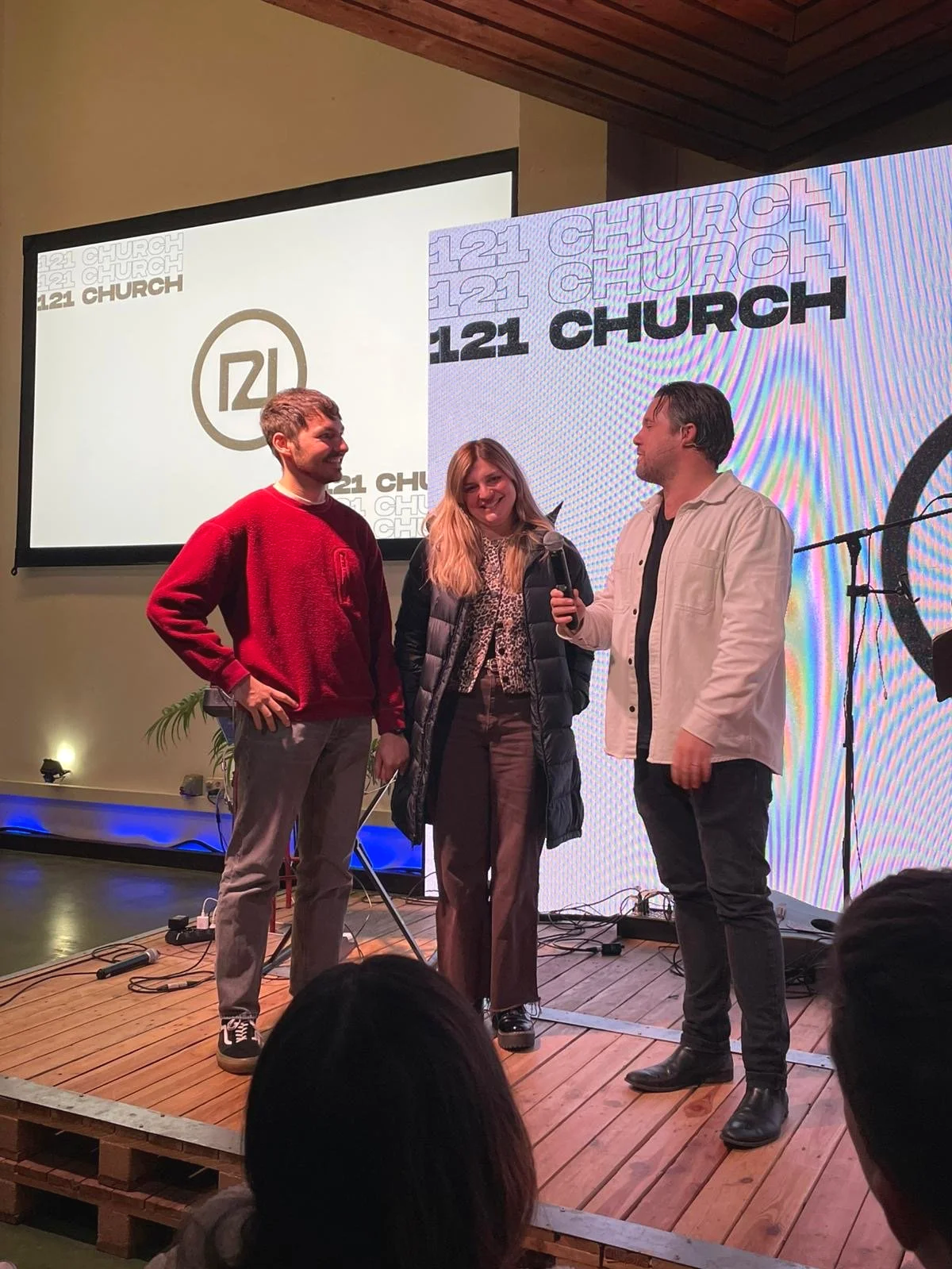 Three people standing on a small wooden stage with microphones, smiling and talking to each other, during a church event called '121 Church'. There is a large screen behind them displaying the event's logo and name.