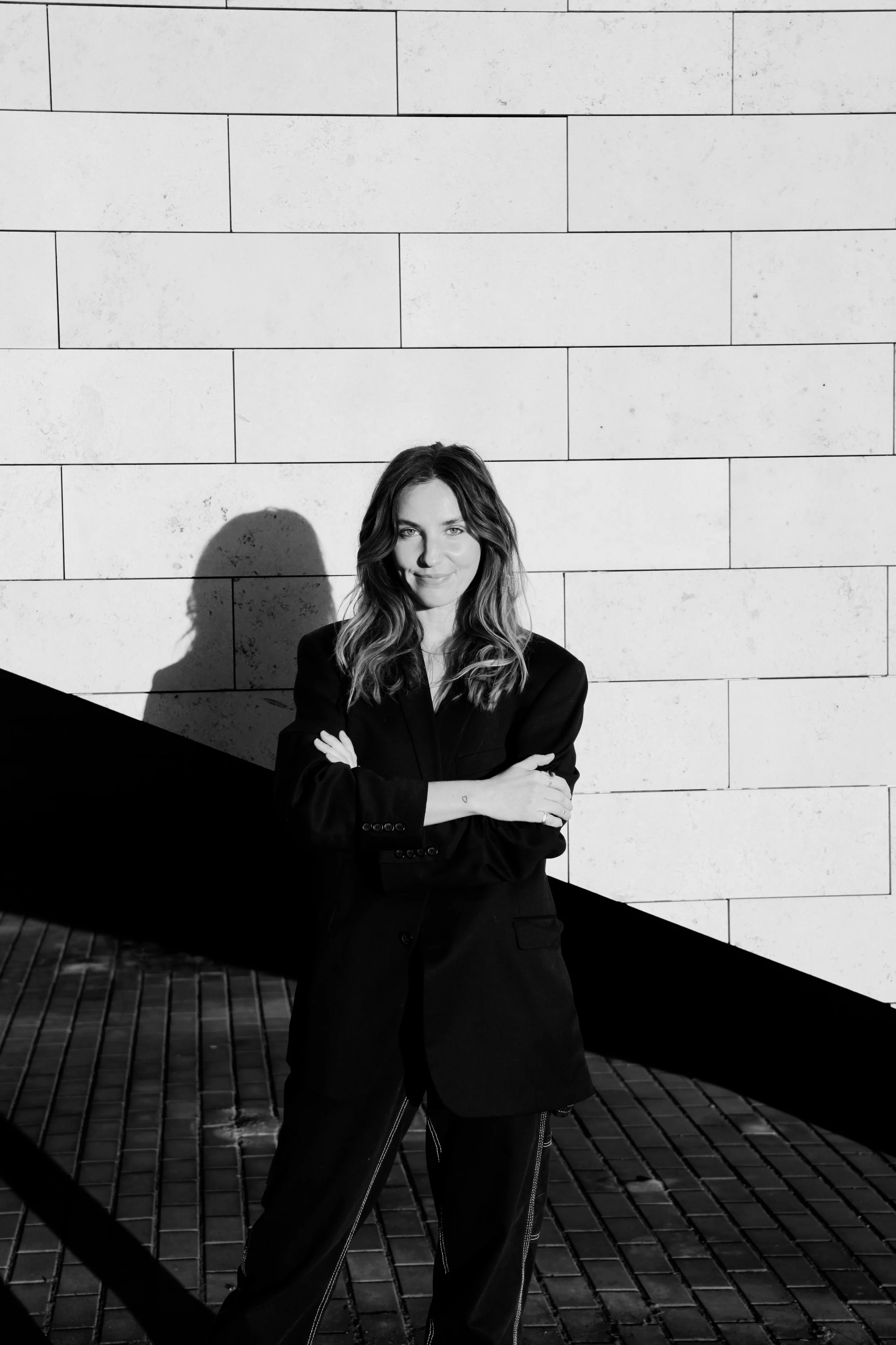 A woman with wavy hair and a confident expression, standing against a light-colored brick wall background. She crosses her arms and is dressed in a dark blazer and pants. The photo is in black and white with shadows cast on the wall.