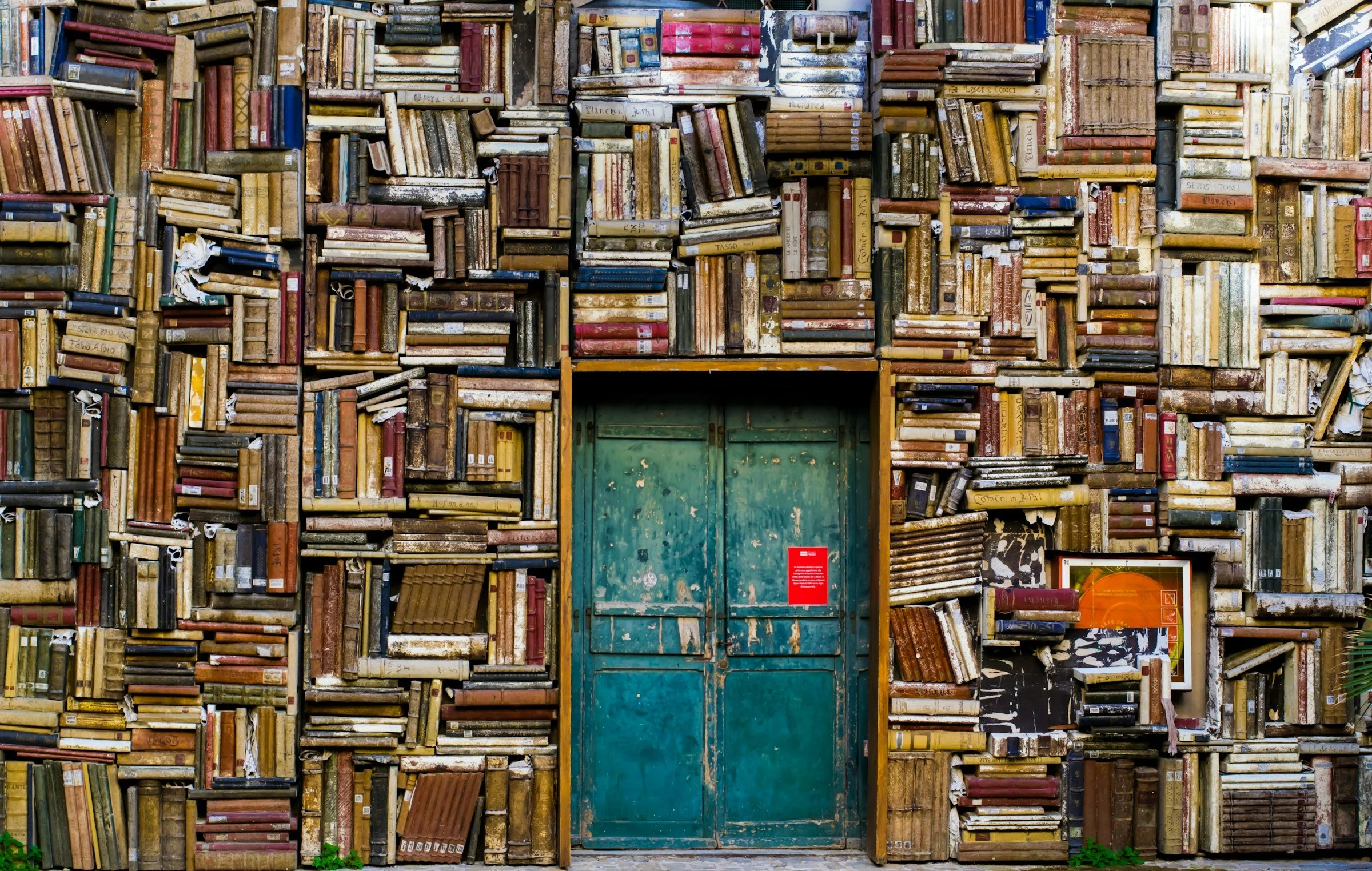 Une façade recouverte de livres anciens empilés de façon désordonnée, avec une porte en métal au centre et un panneau rouge à son dessous.