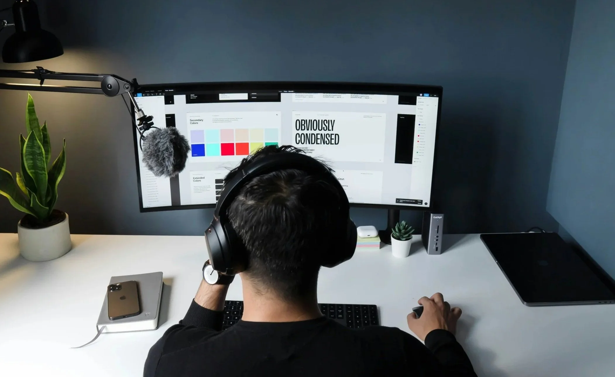 Une personne portant un casque d'écoute assise devant un ordinateur à double écran, travaillant ou regardant l'écran, avec une plante, un téléphone portable et une petite plante en pot sur le bureau.