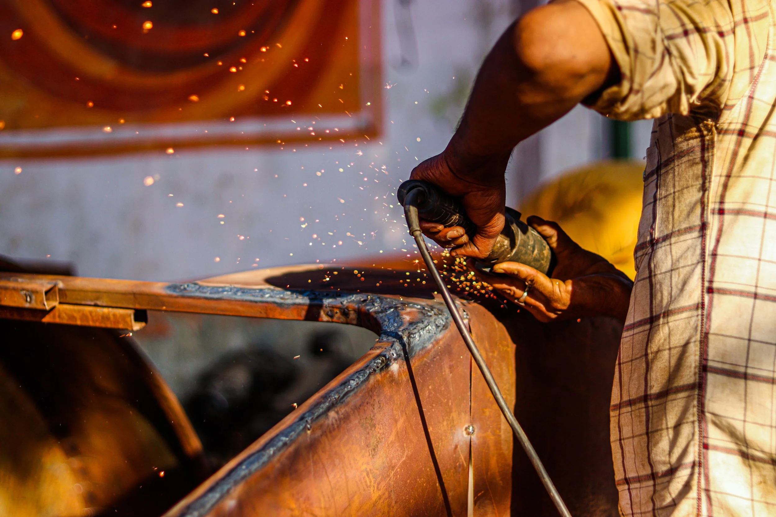 A worker welding a metal surface, sparks flying, wearing a short-sleeved shirt with a plaid pattern.