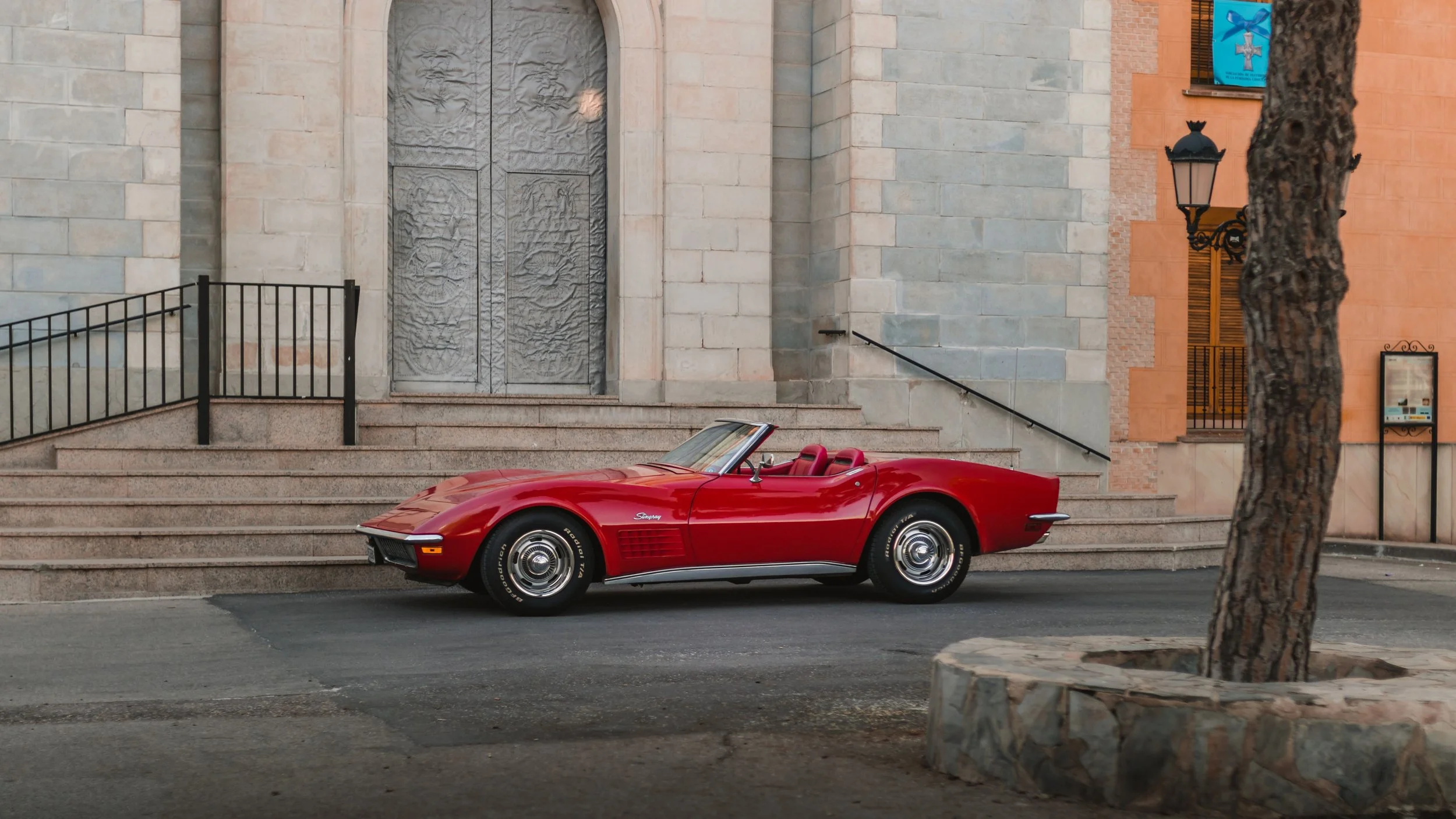 A red vintage Chevrolet Corvette convertible car parked on a city street in front of a building with stone steps and a large metal door.