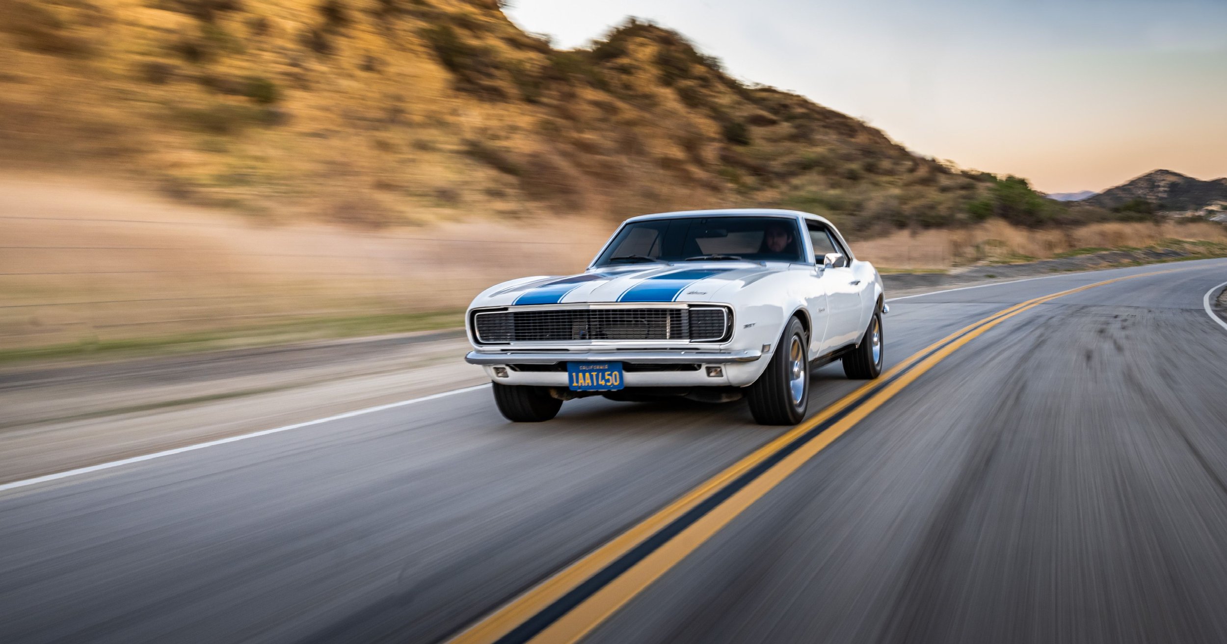 A white vintage muscle car with blue racing stripes driving on a winding mountain road, kicking up dust behind it during sunset.