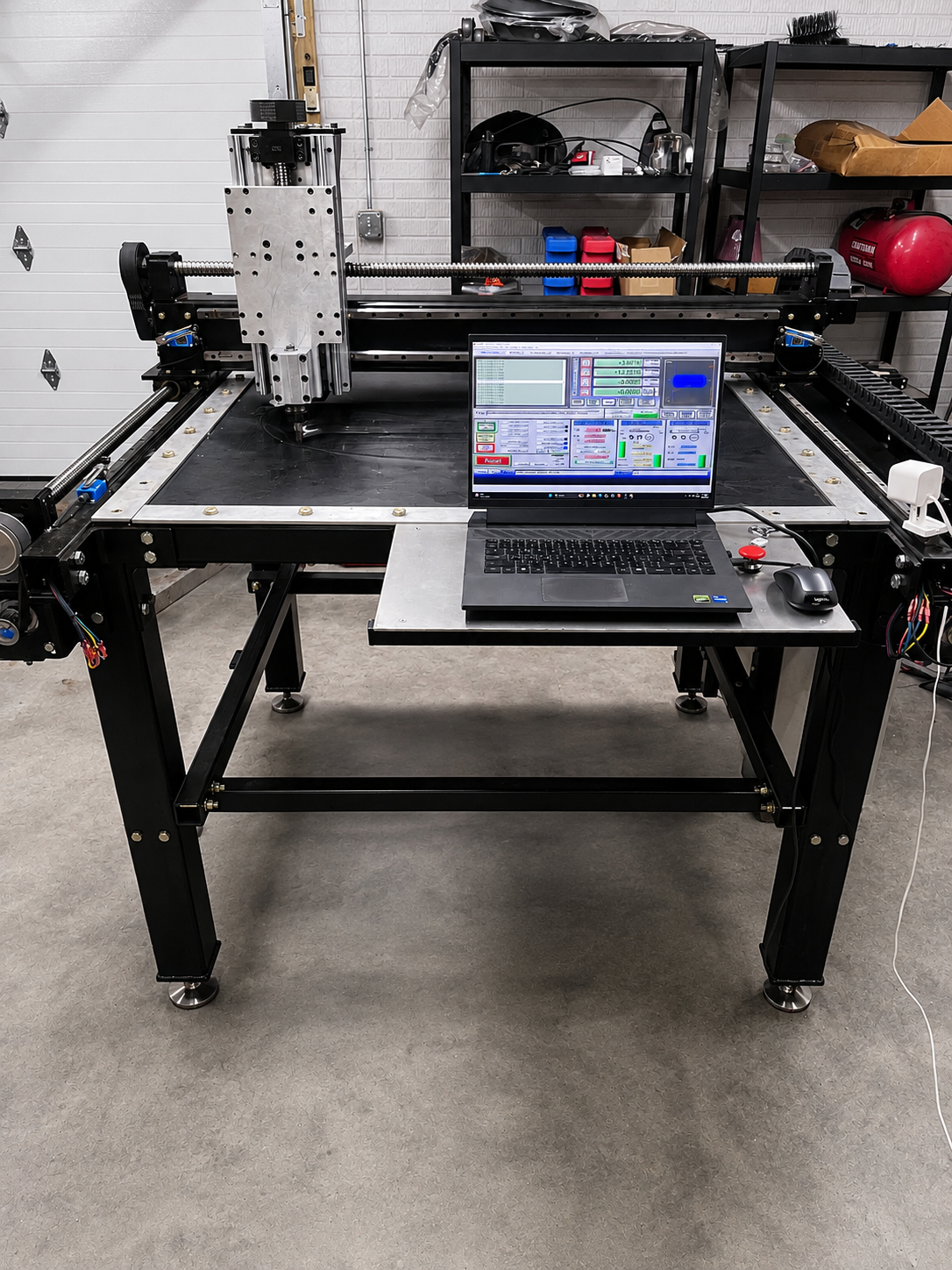 A CNC machine in a workshop with a laptop and mouse on the table, and a computer screen displaying machine controls.