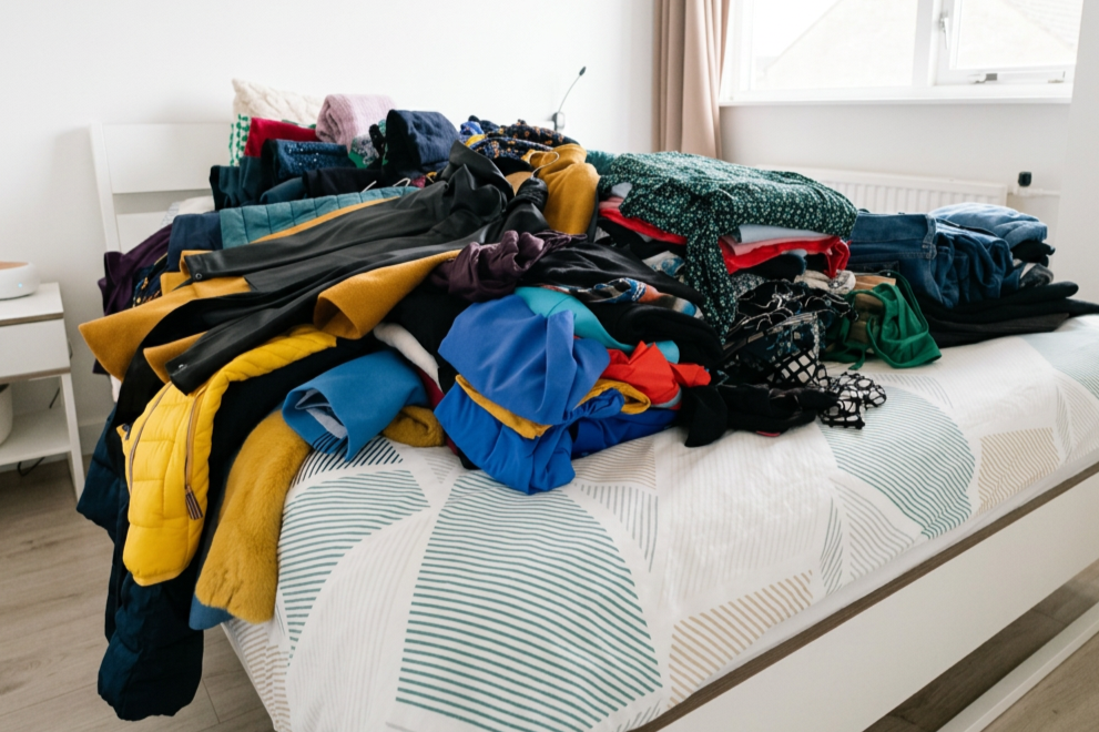 An unmade bed with a pile of assorted clothes and textiles on top in a bedroom with natural light from a window.
