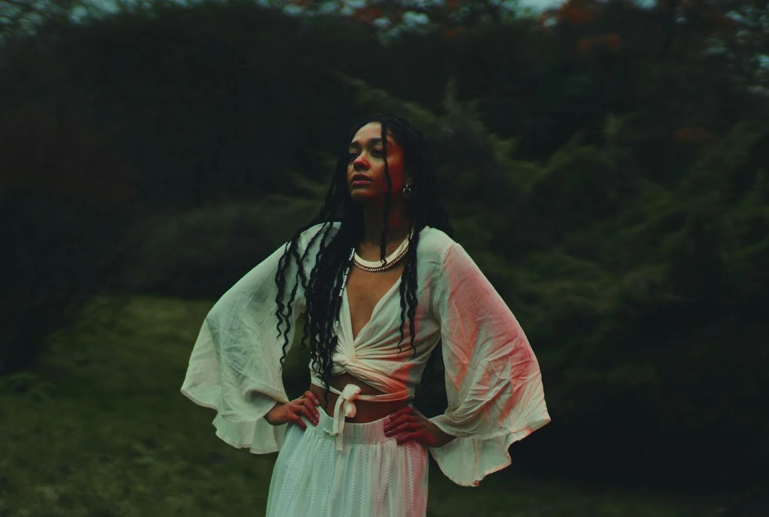 A woman with long dreadlocks standing outdoors with a hand on her hip, wearing a white tied-up blouse and matching flowy pants, with mountains and greenery in the background.