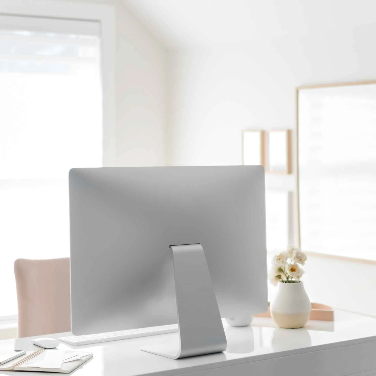 Apple computer on a white desk with a pink chair, a vase of white flowers, and a window in the background.
