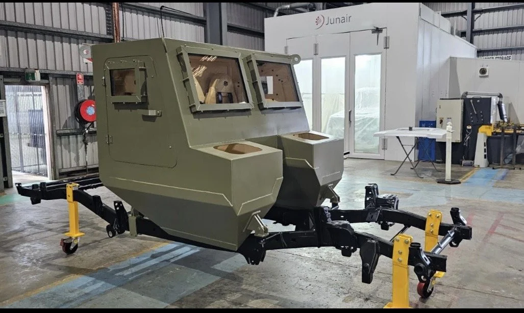 An armored vehicle simulator or training pod with a military design, equipped with windows and mounted on a wheeled black and yellow chassis inside a warehouse or industrial facility.