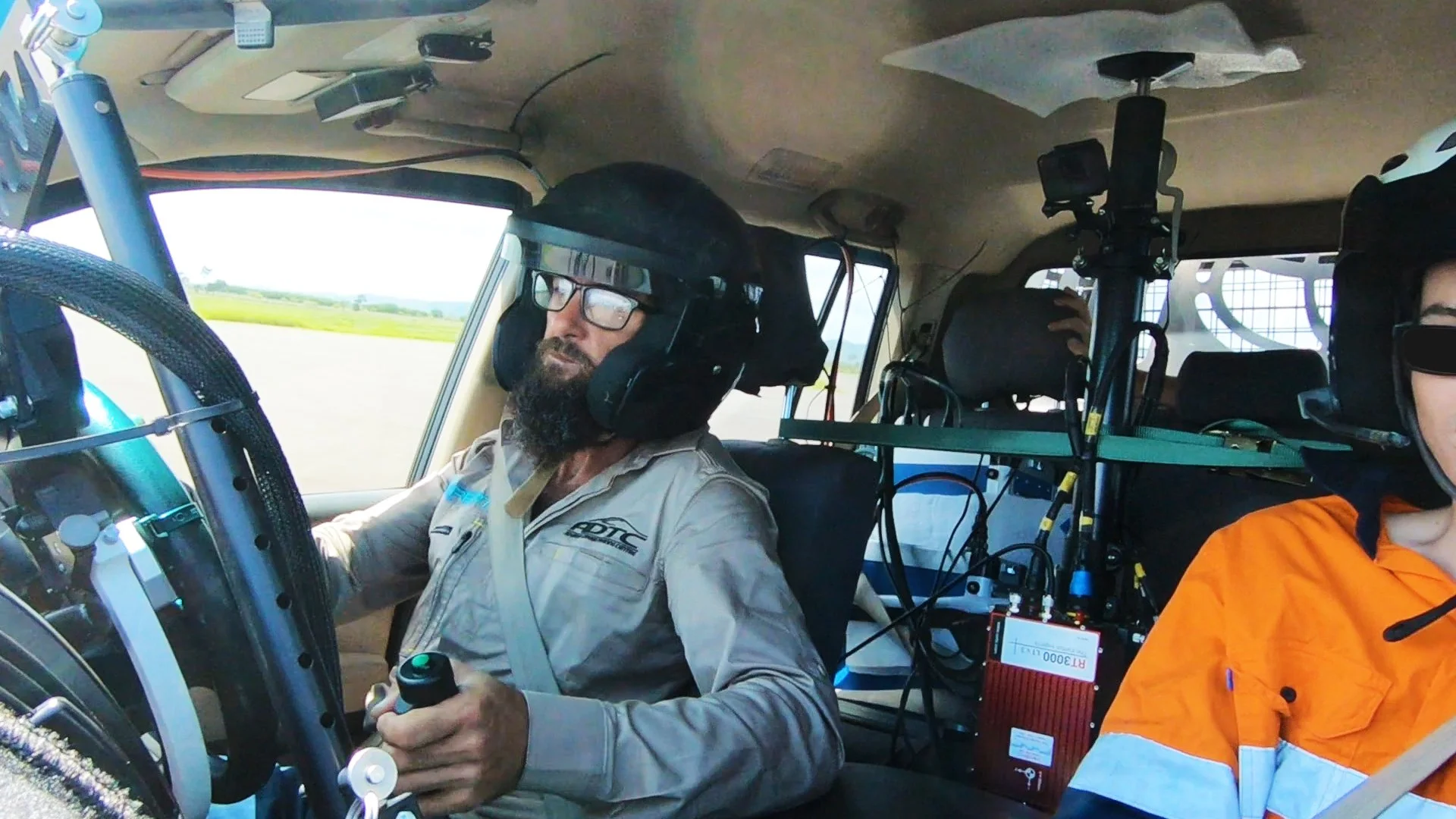 Two researchers in a helicopter, wearing helmets with headsets and goggles, surrounded by electronic instruments and wires, flying over a rural landscape.