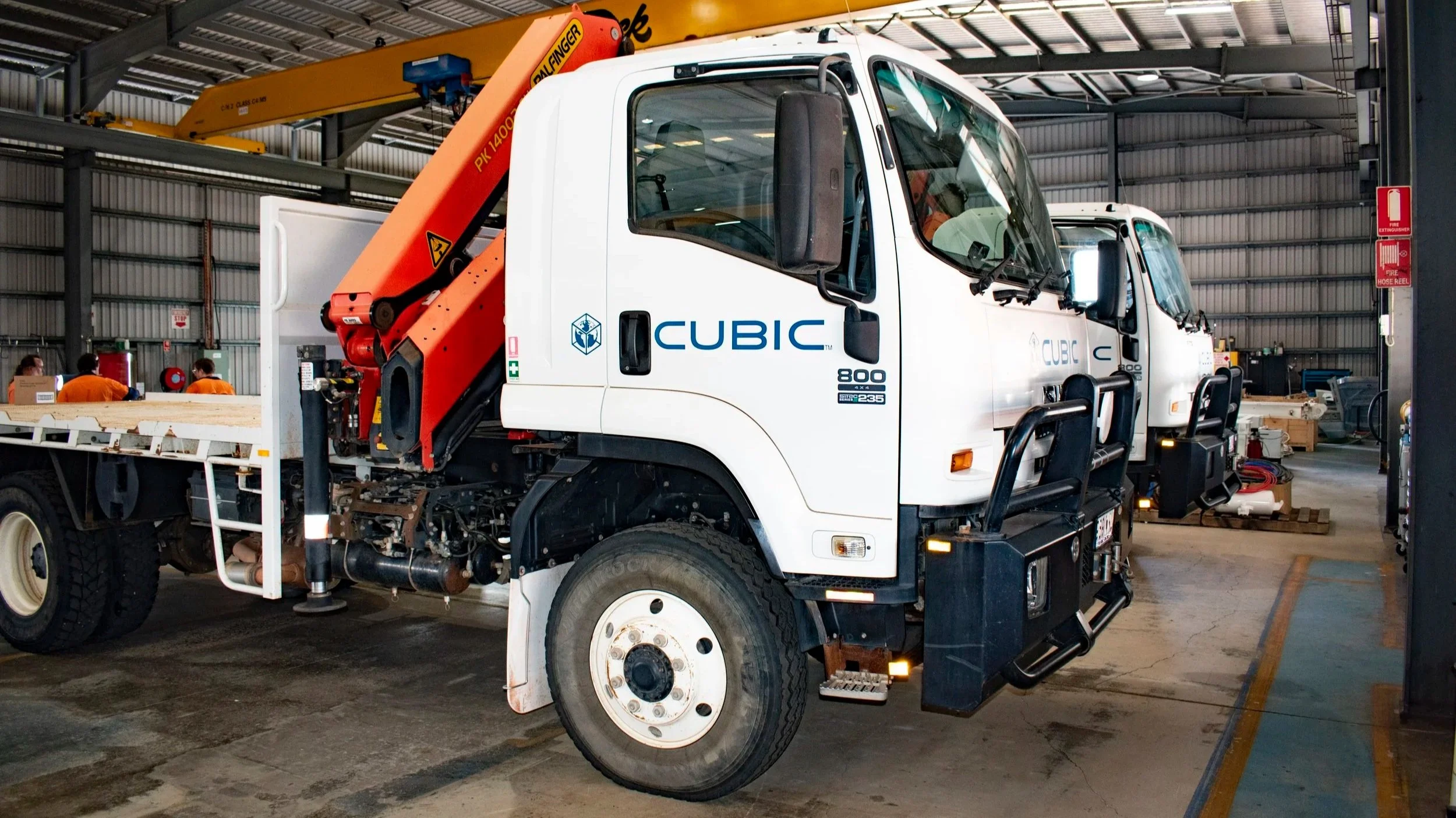 Indoor warehouse with a white utility truck labeled 'CUBIC' and equipped with a crane arm, surrounded by workers, equipment, and storage items.