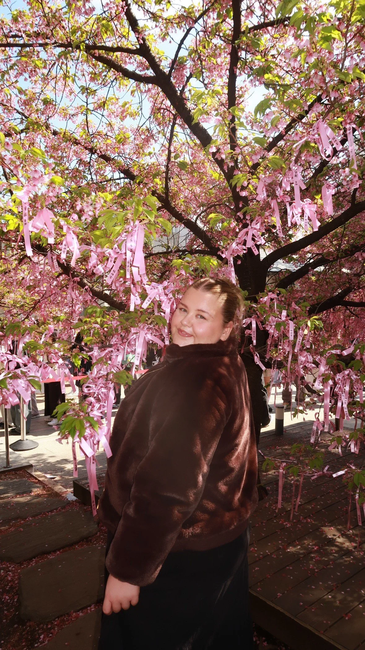 A woman standing in front of a pink cherry blossom tree decorated with pink ribbons. The woman is smiling and wearing a dark brown jacket, and the sun is shining.