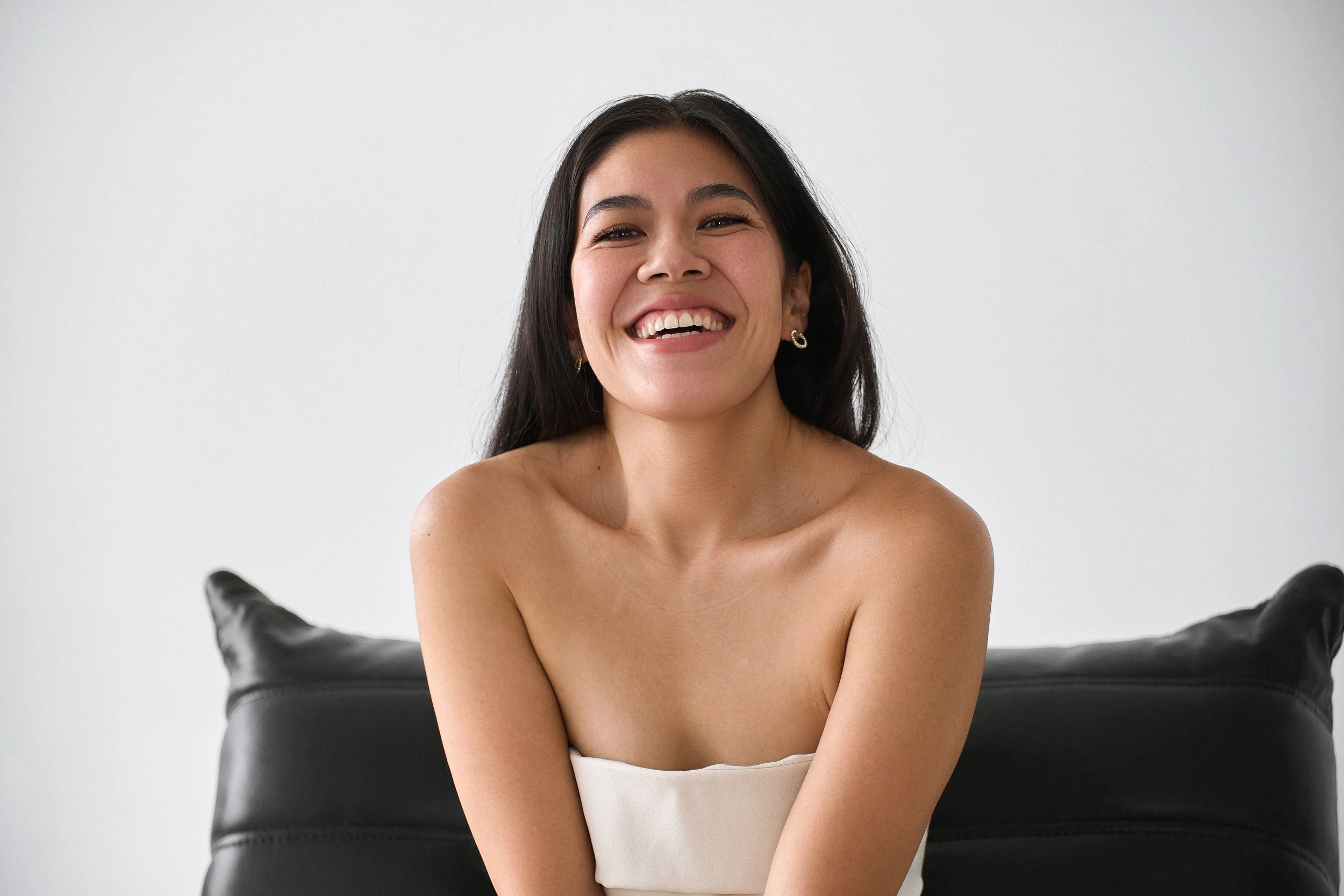 A smiling woman with long dark hair, wearing earrings, sitting on a black couch in front of a plain white wall.