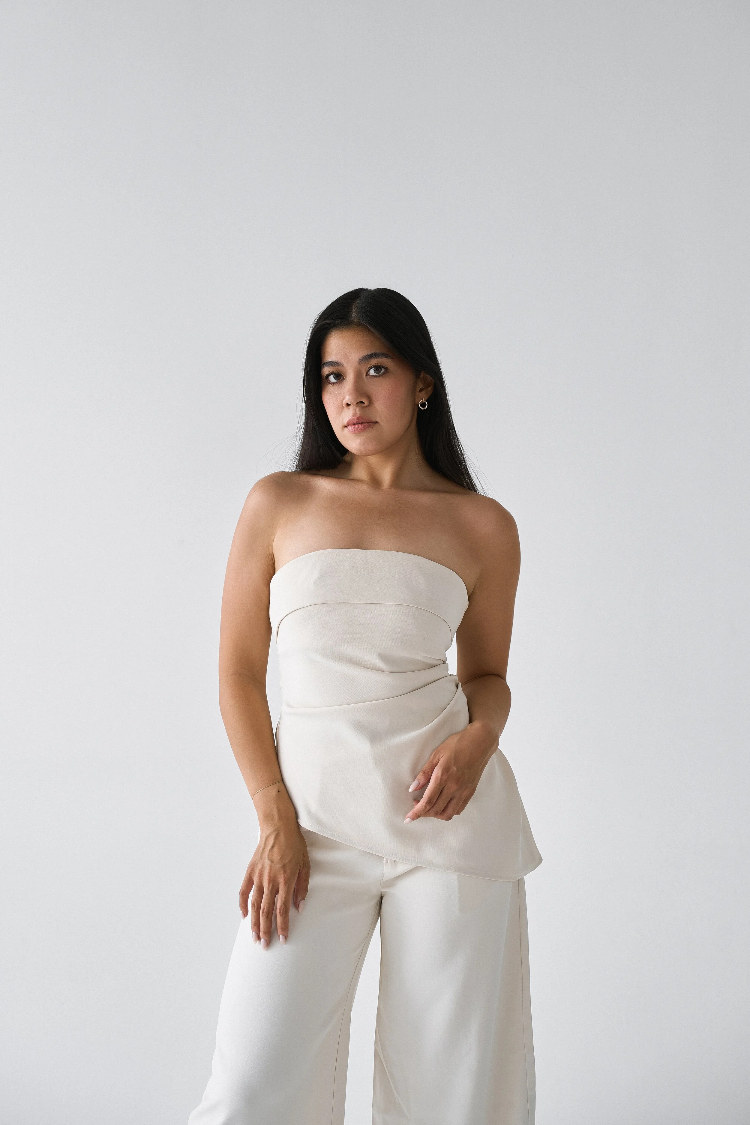 A woman with dark hair wearing a strapless white top and matching white wide-leg pants against a plain light gray background.