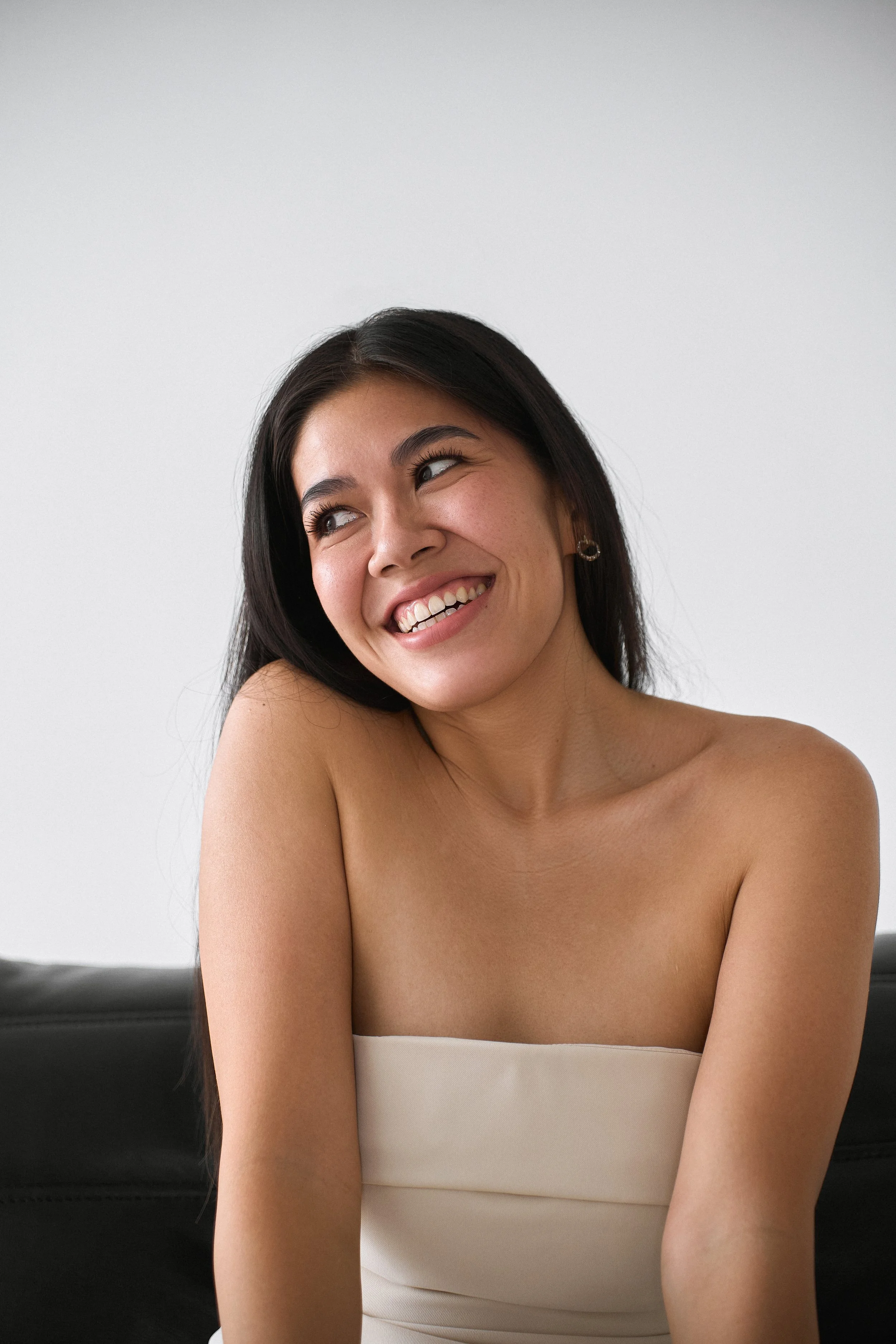 A young woman with long dark hair is smiling and looking to her left. She has a tan skin tone, is wearing hoop earrings, and a white strapless top. She is seated in front of a white wall with a black piece of furniture behind her.