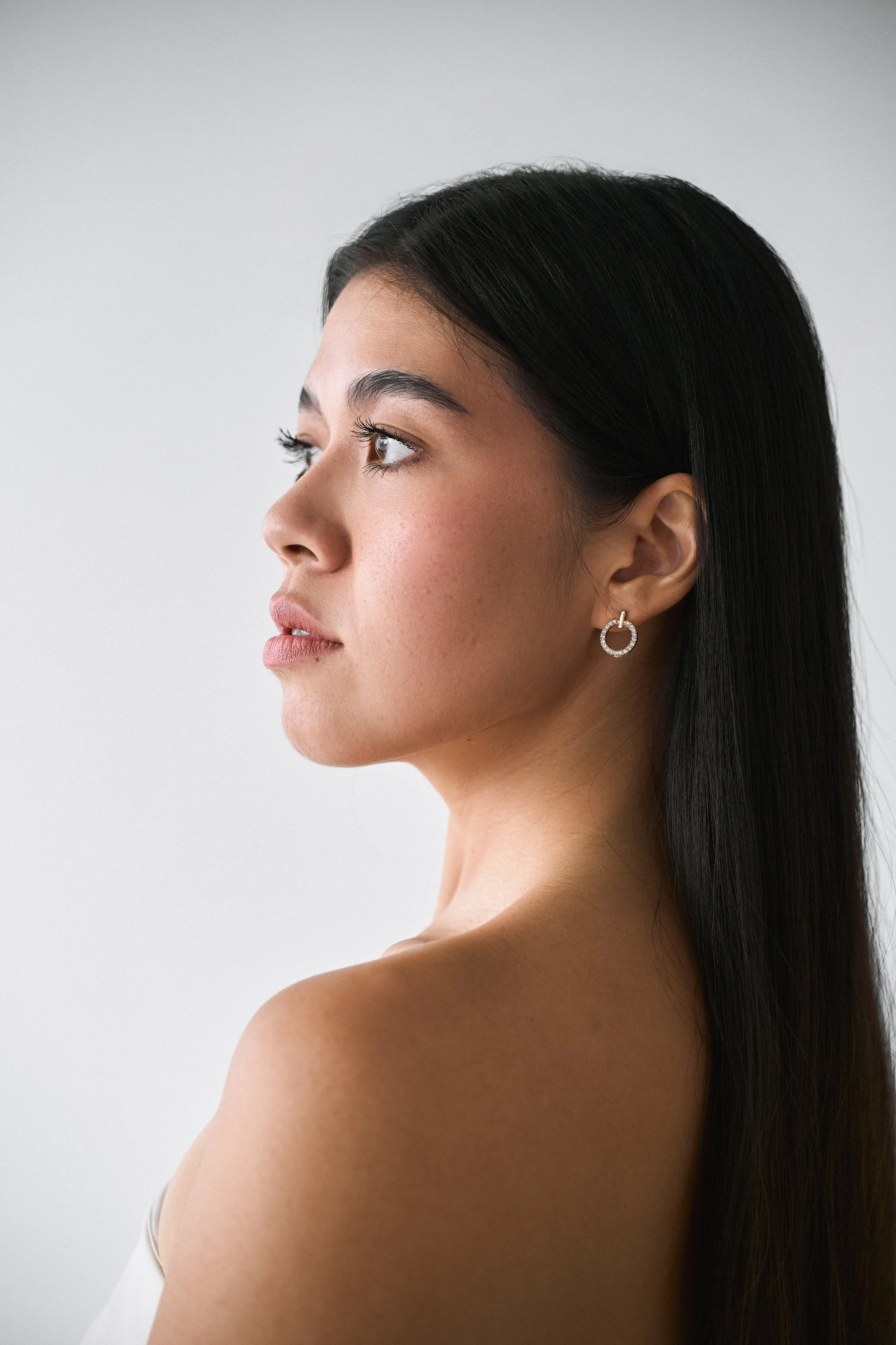 Profile of a woman with long black hair, light skin, wearing hoop earrings, looking to the left, against a plain light background.
