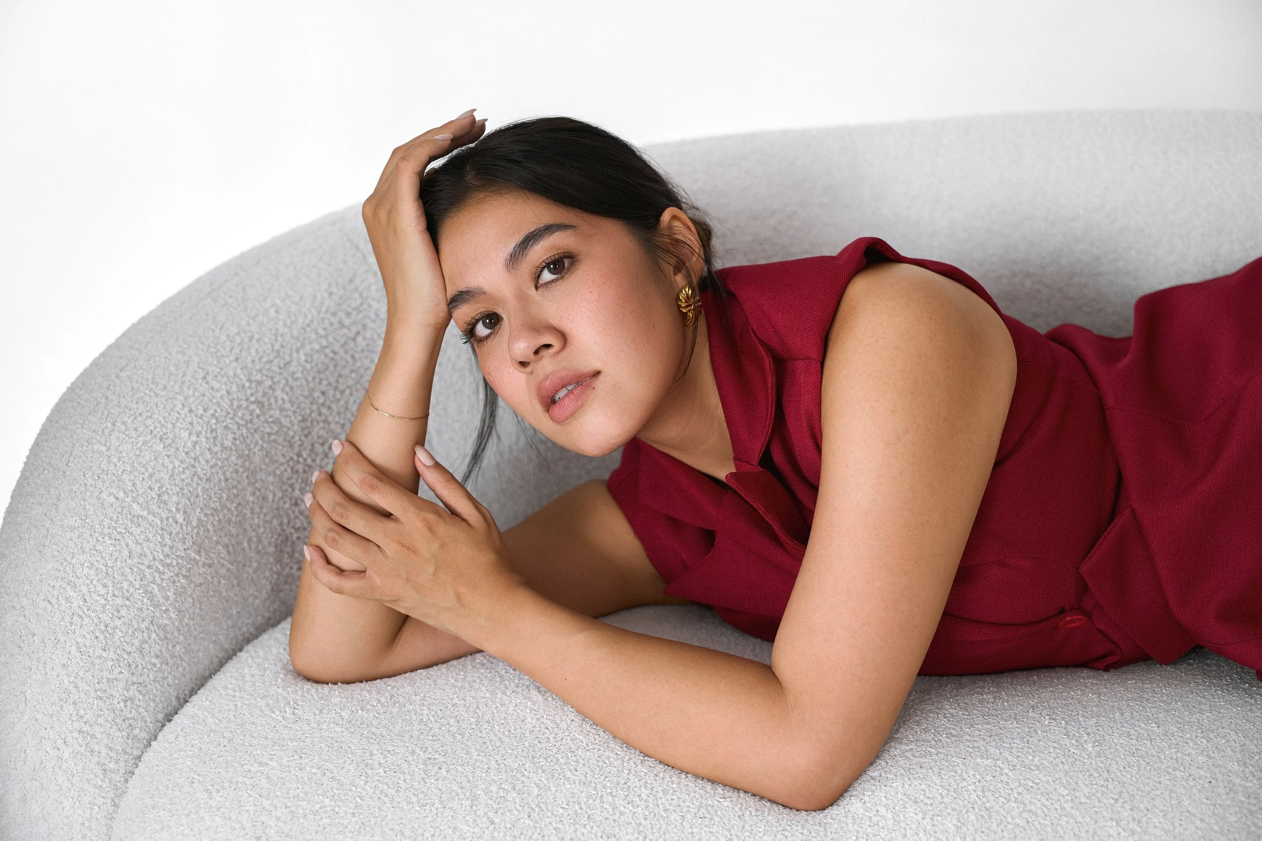 A woman in a red dress reclining on a light-colored textured sofa, resting her head on her hand, looking directly at the camera.