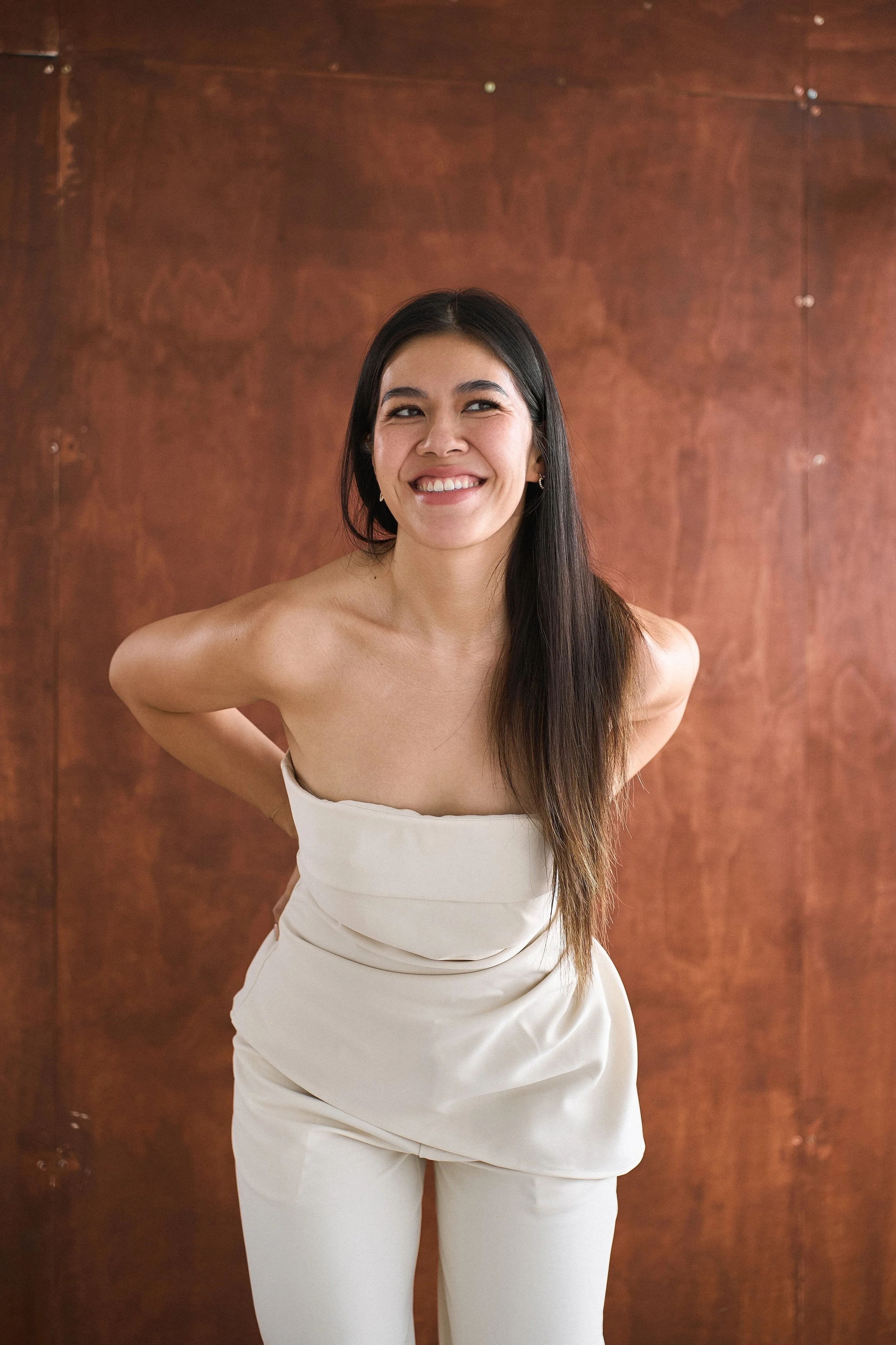 A young woman with long dark hair smiling and wearing a strapless white dress. She stands in front of a wooden background with her hands behind her back.
