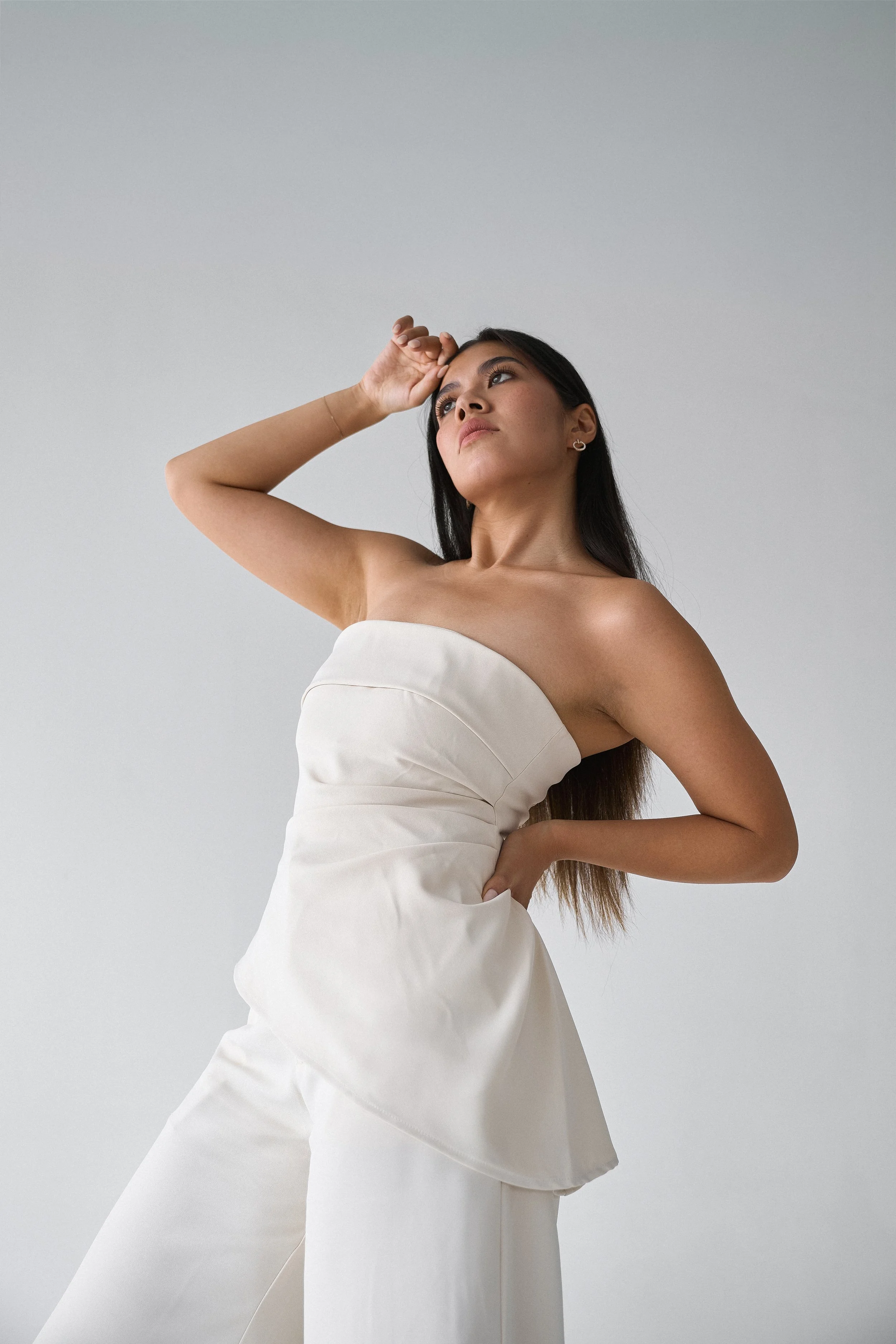 A woman with long dark hair in a strapless white dress poses against a plain gray background. She has one hand resting on her forehead and looks off to the side.