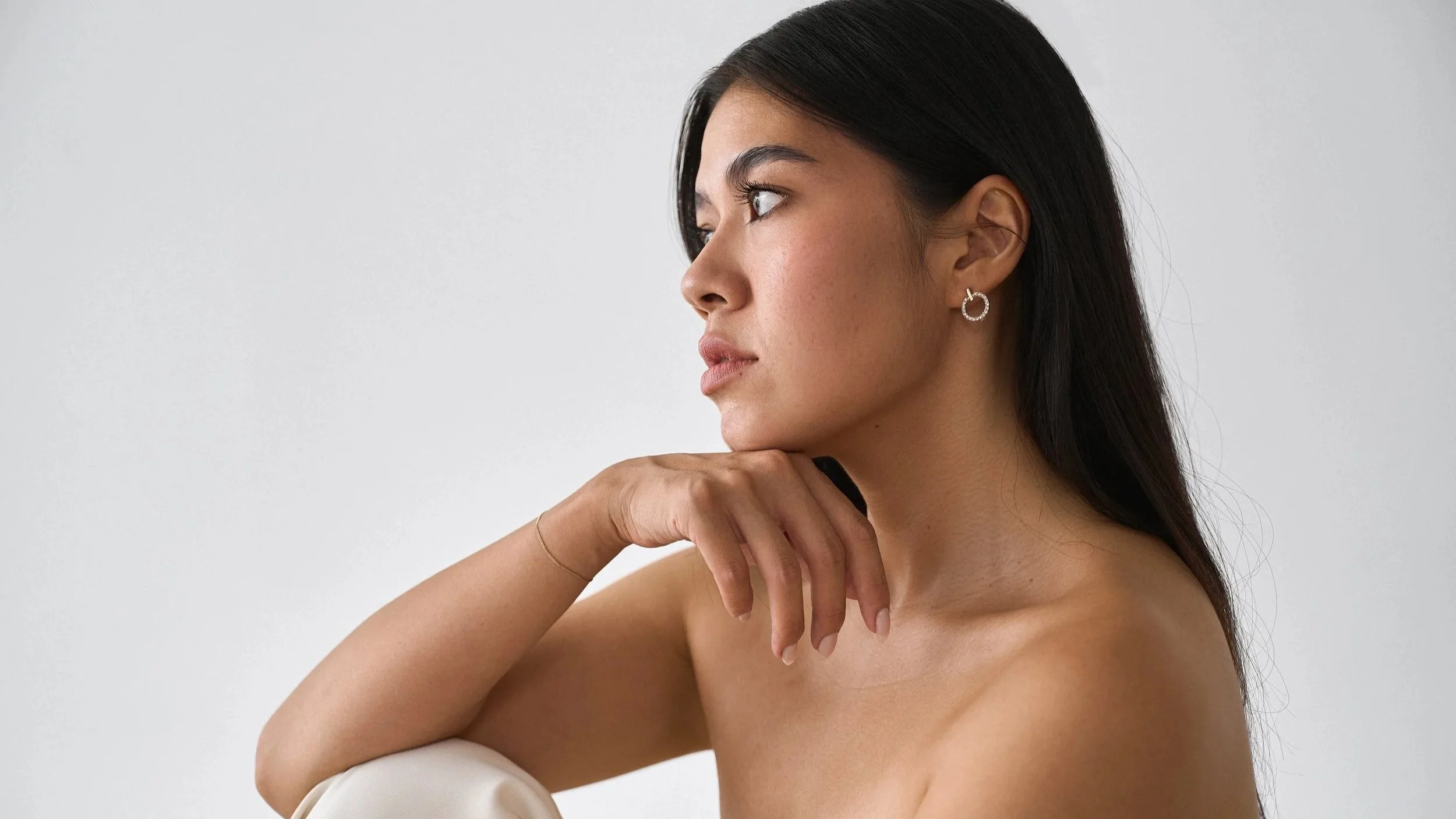 Profile view of a woman with long dark hair, wearing hoop earrings, resting her chin on her hand, against a plain light background.