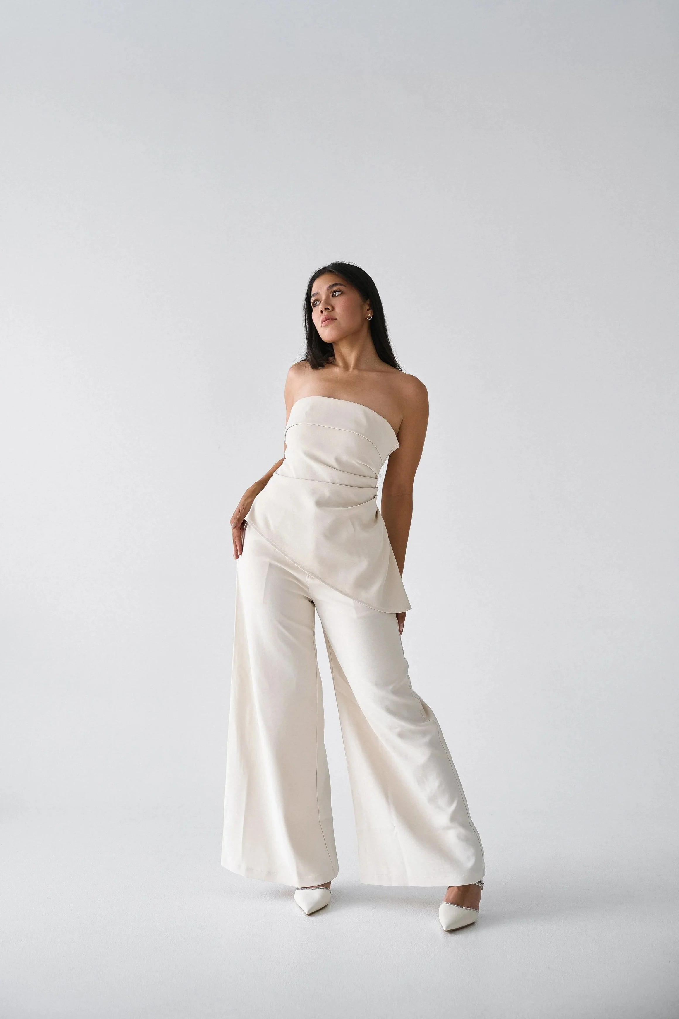 A woman in a strapless cream-colored jumpsuit with wide-leg pants standing against a plain light gray background.