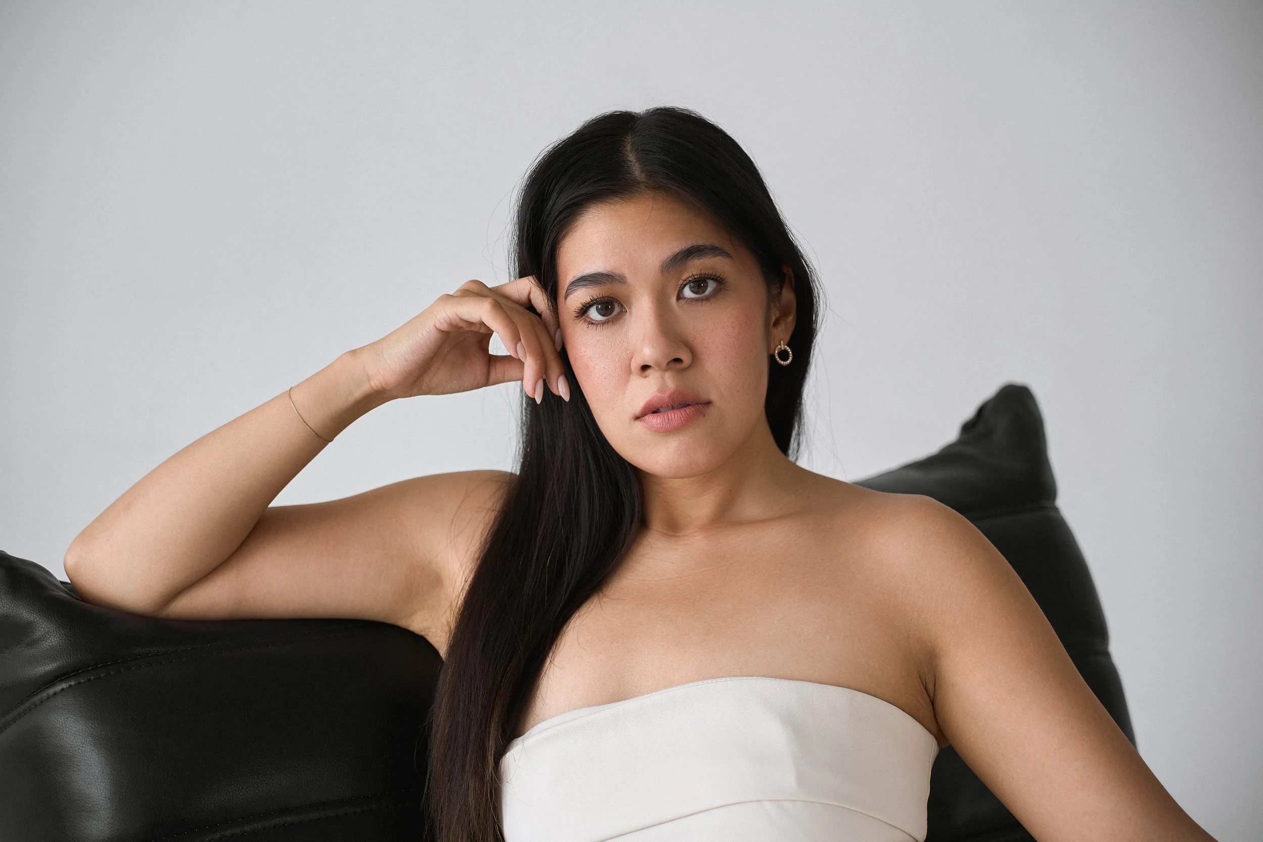 A young woman with long dark hair, wearing earrings, lying on a black leather couch, wearing a white strapless top, resting her head on her hand, with a neutral expression against a plain light gray wall.