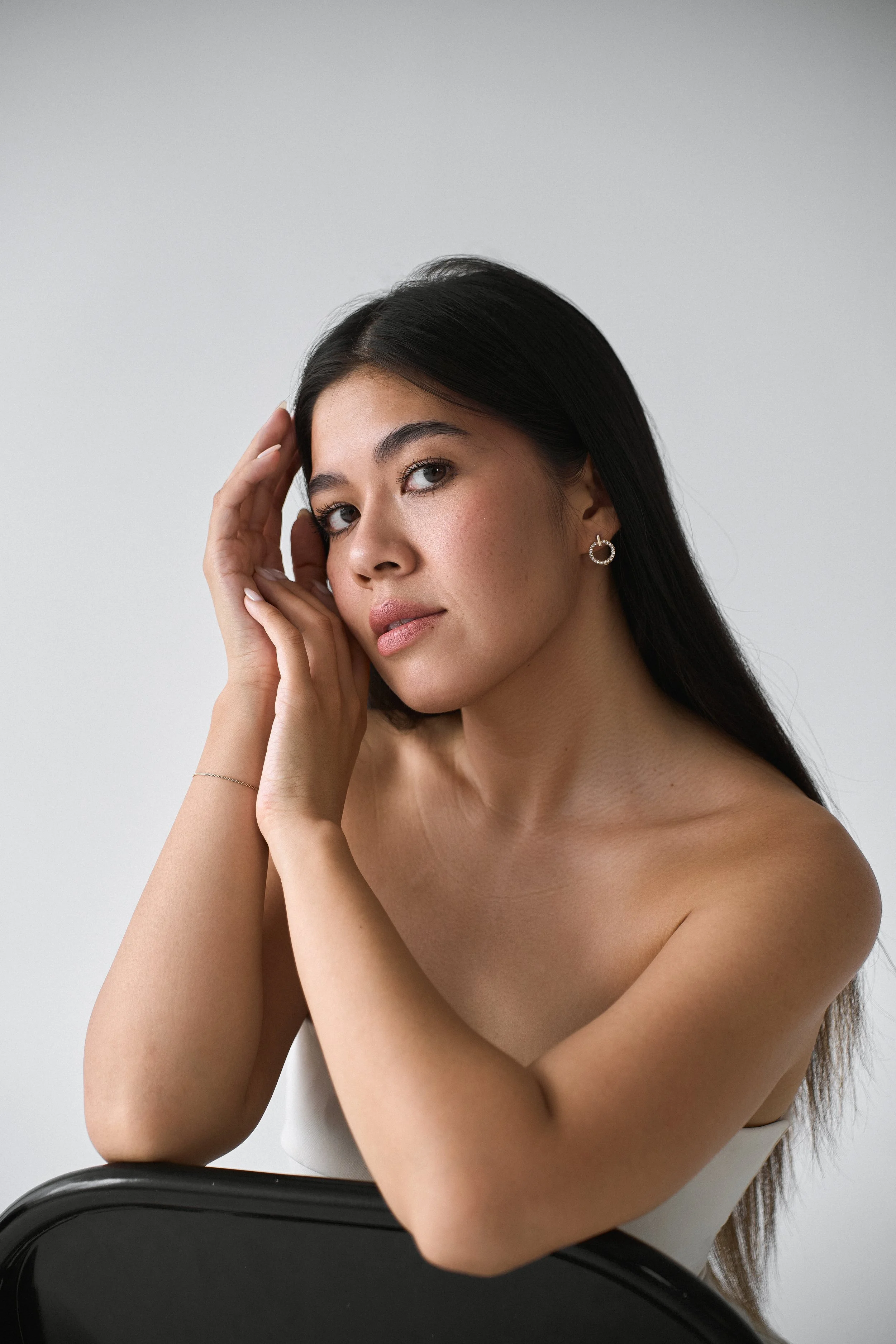 A woman with long dark hair and earrings sitting on a chair, topless with her arms crossed in front of her, against a neutral background.