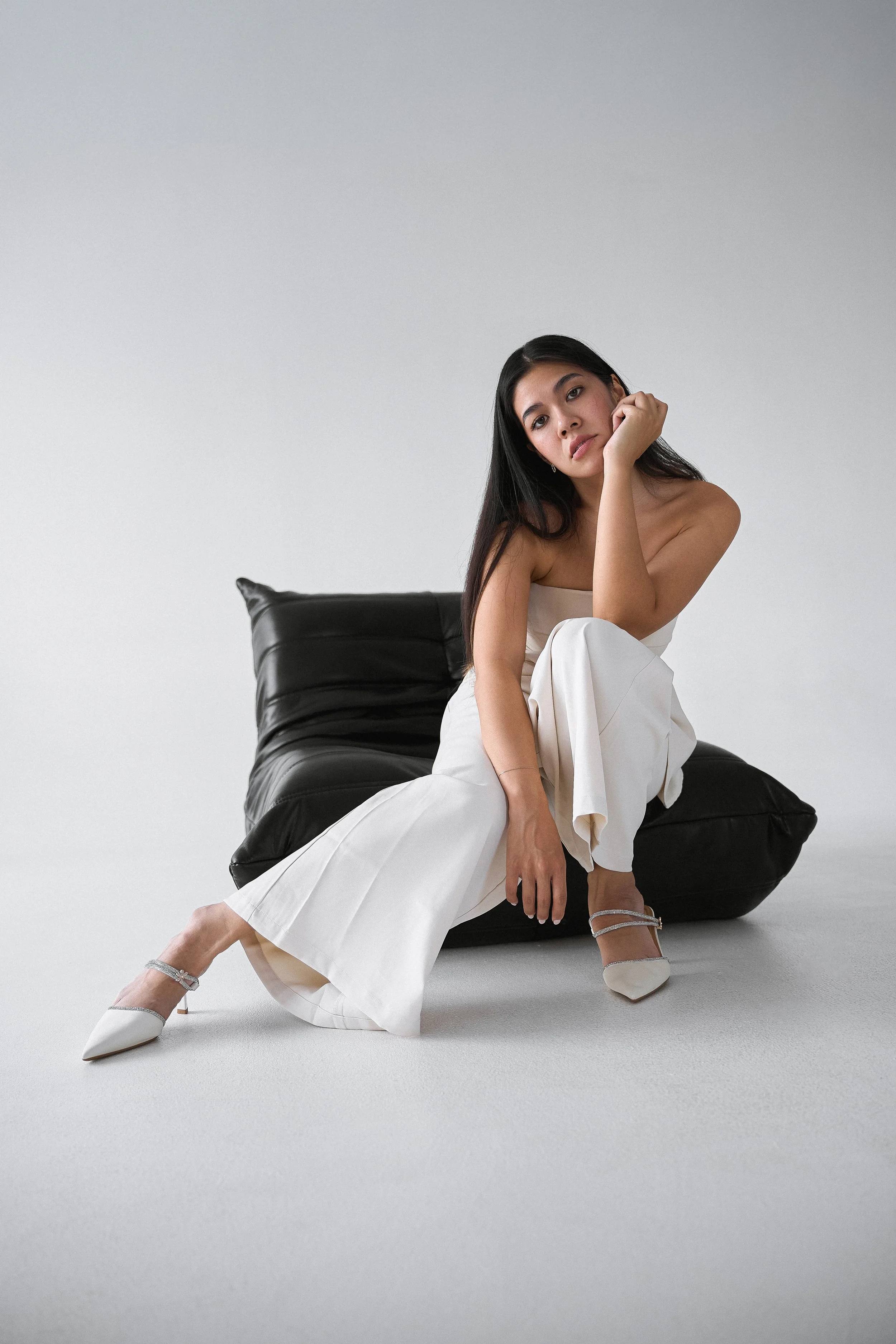 A woman sitting on the floor next to a black sofa, dressed in white pants and high heels, posing with her head resting on her hand in a minimalistic studio setting.