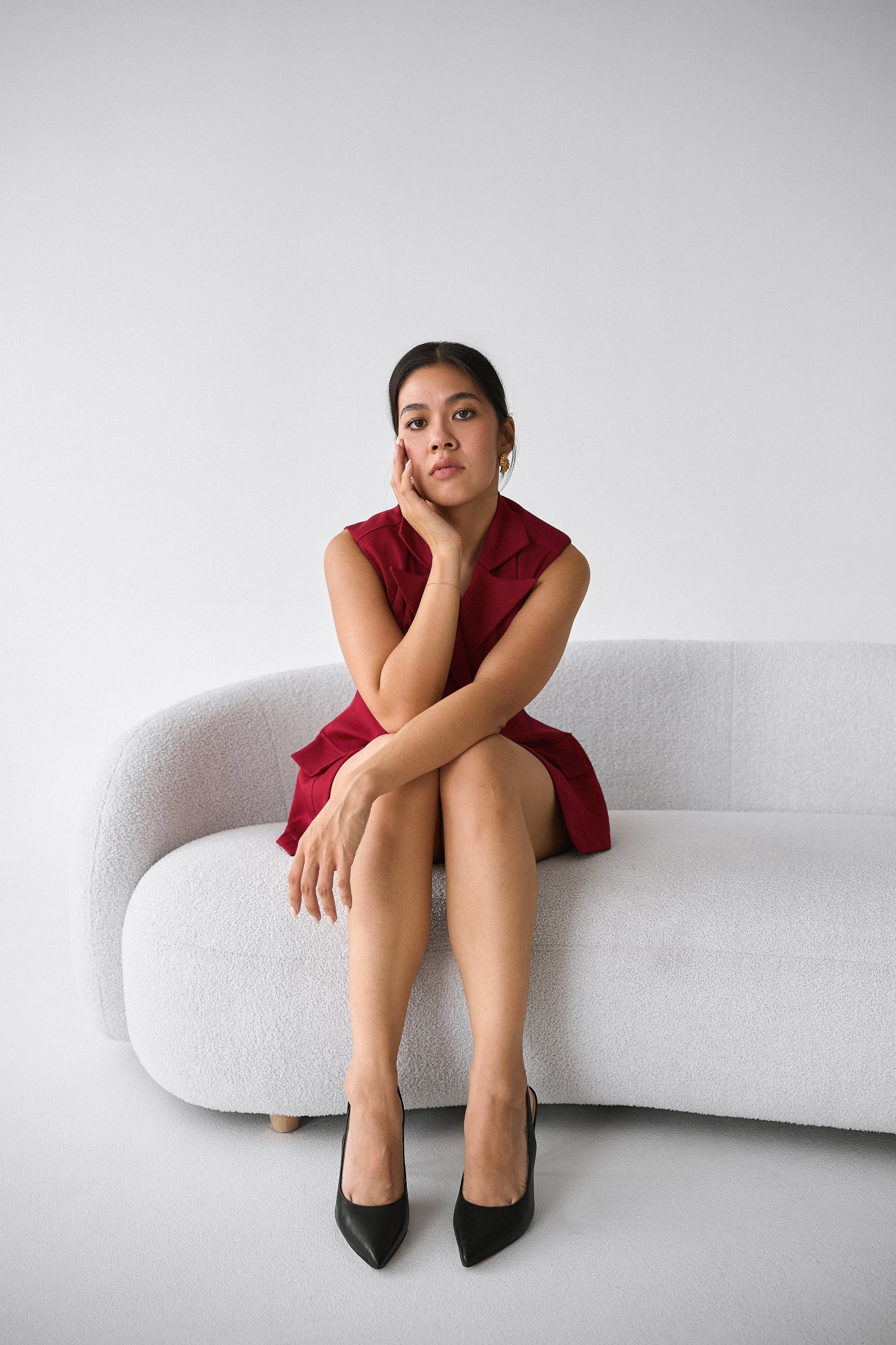 A woman in a red dress sitting on a white textured sofa with her hand on her face, looking directly at the camera, with high heels on, in a minimalist setting.