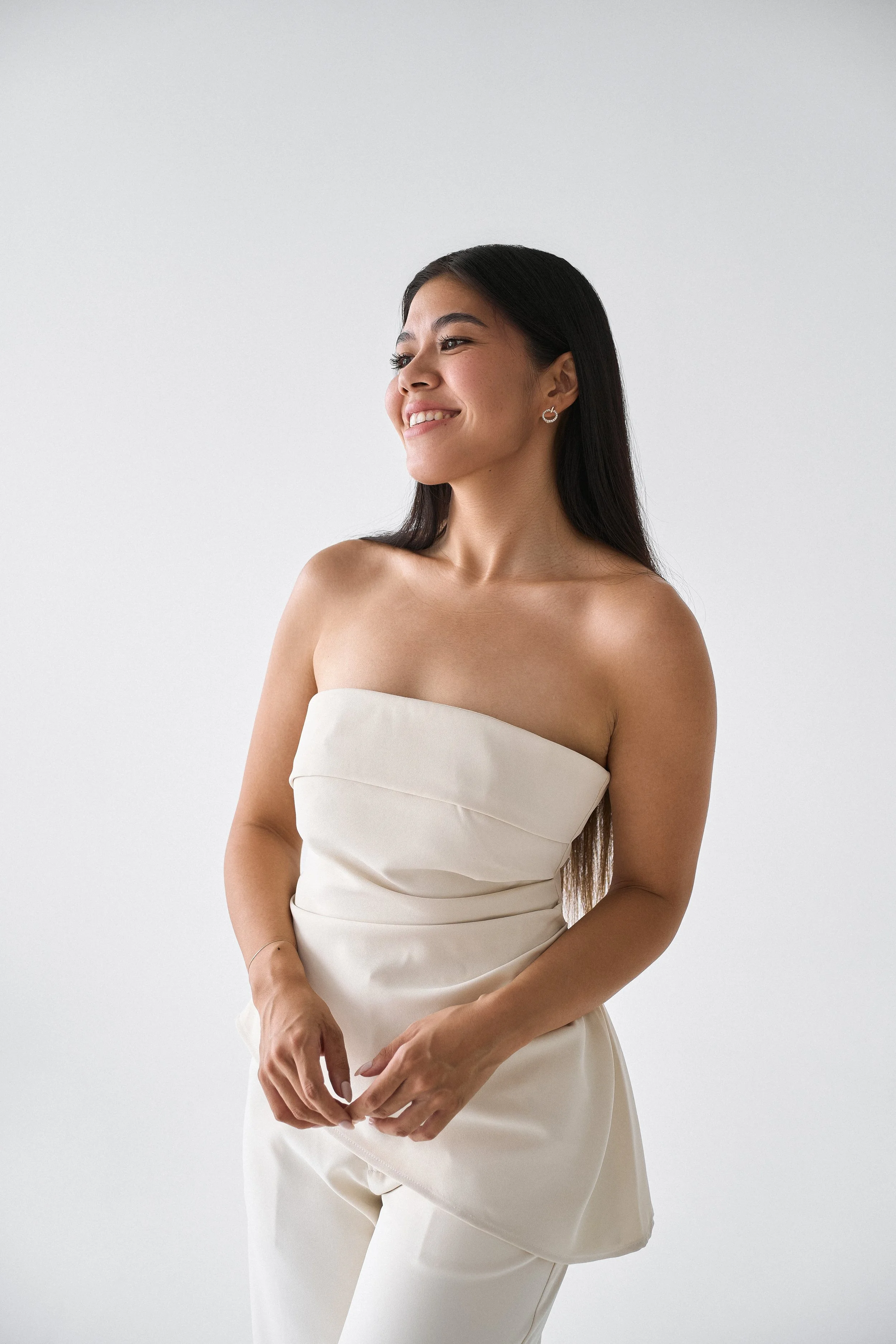 A woman with long dark hair, wearing a strapless cream-colored top and matching pants, standing against a plain white background, smiling slightly and looking to the side.