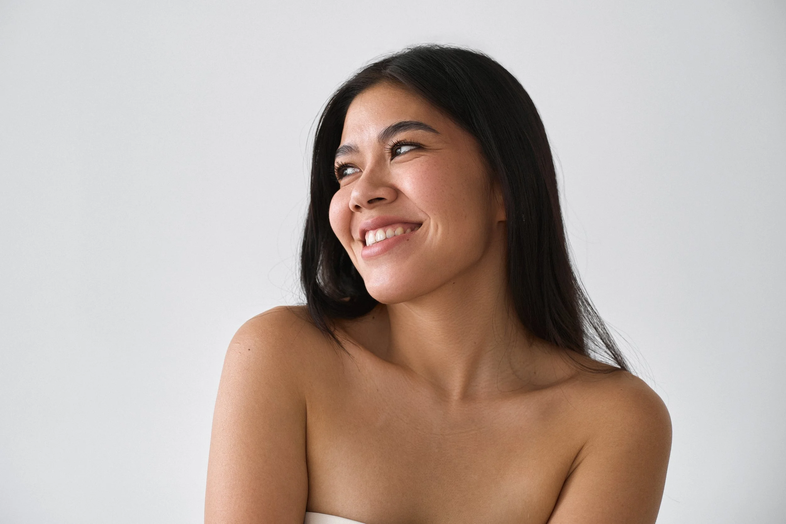 Close-up of a woman with long dark hair smiling and looking to the side, against a plain light gray background.