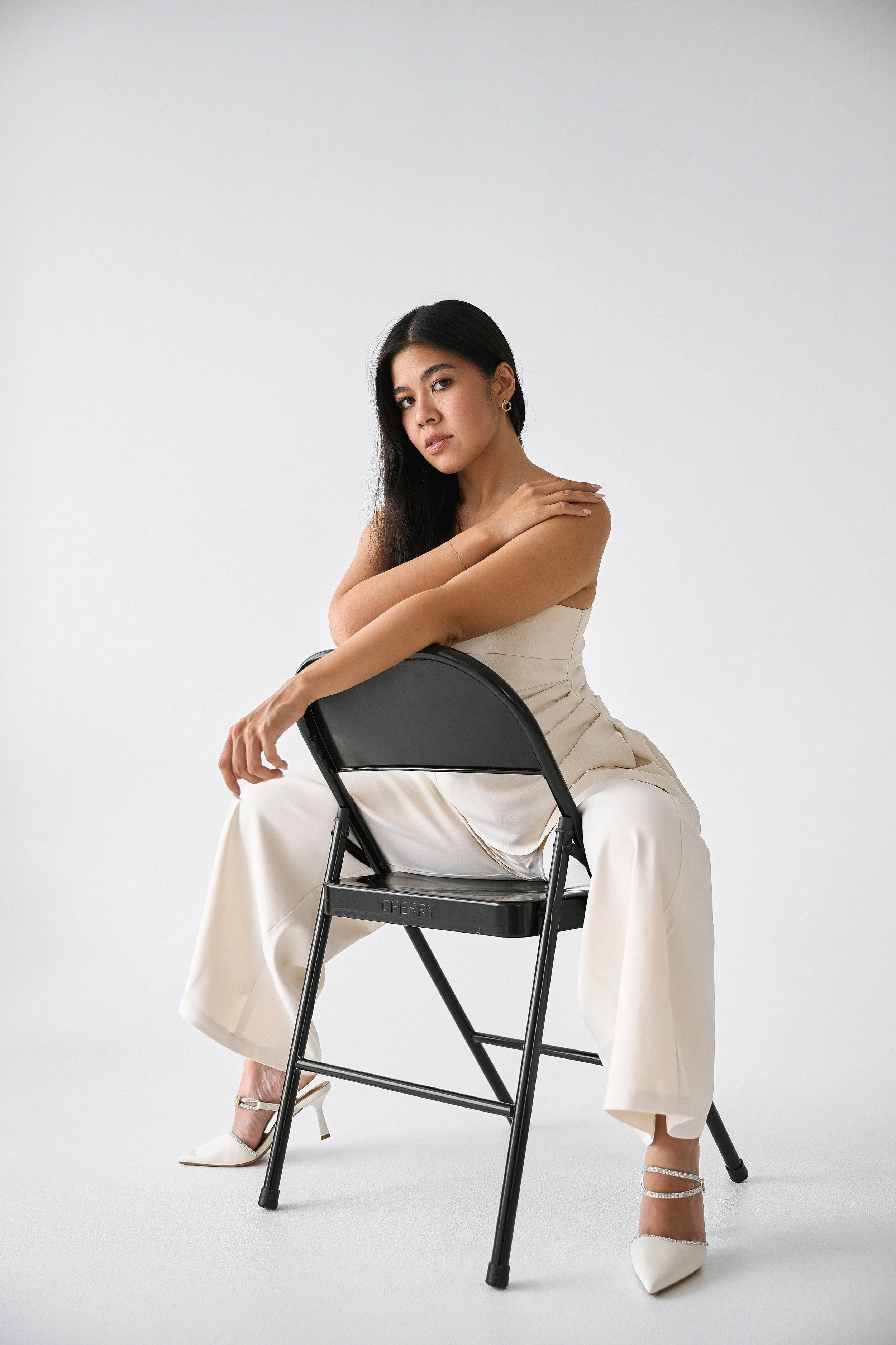 A woman sitting on a black folding chair, dressed in beige pants and a beige sleeveless top, with her arms crossed over her shoulder. She is wearing white pointed-toe heels with ankle straps, and has long black hair. The background is plain light gra